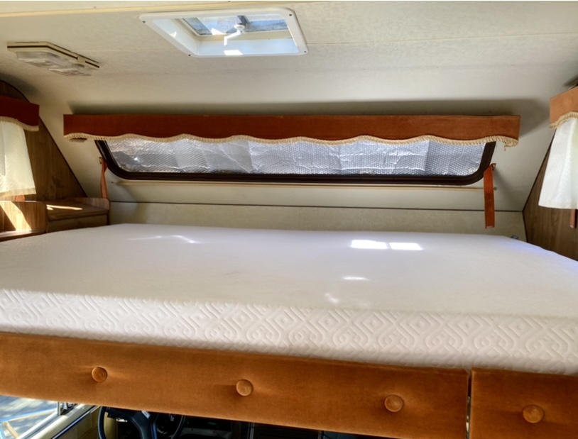 Inside an RV rental: a neat cab-over bunk with a white mattress, window sunshade, and retro valances.. Gmc Motorhome 1985