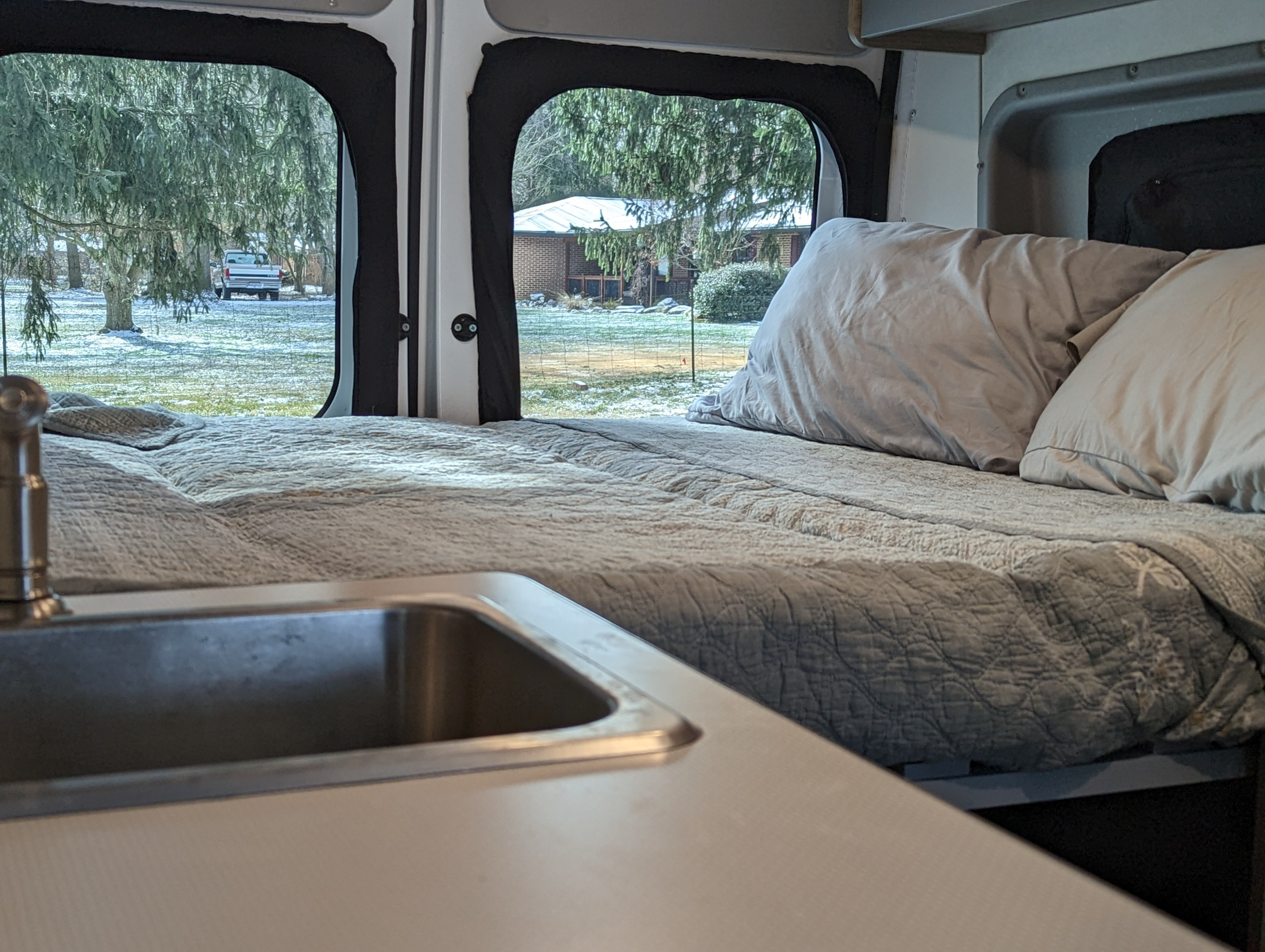 Inside a cozy RV rental: a comfortable bed and sink with a snowy outdoor view of trees, truck, and house.. Winnebago Solis 2021
