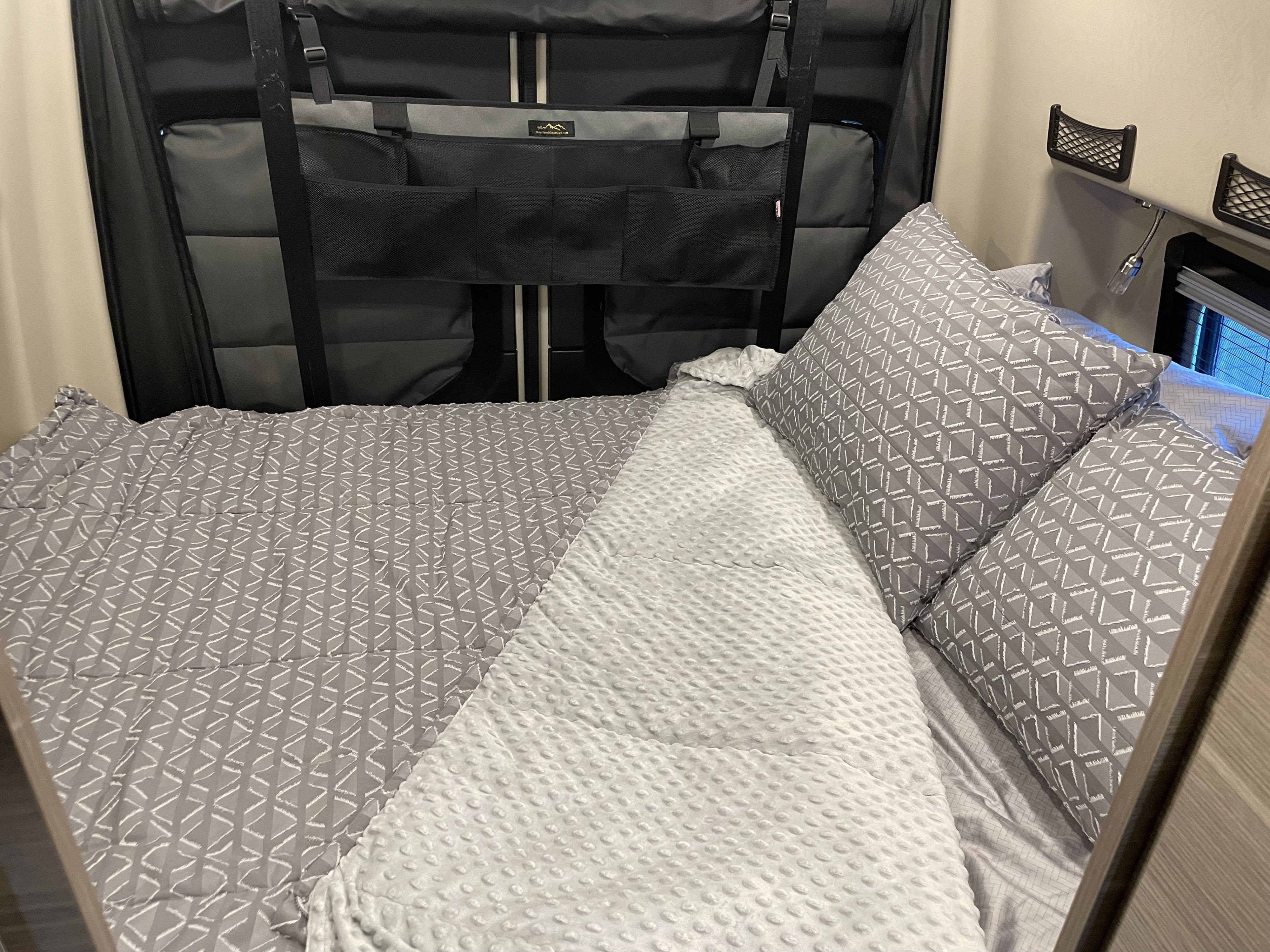 Cozy RV rental bed with patterned gray bedding, cozy minky blanket, and wall organizer for practical storage in a compact space.. Entegra Launch 19Y 2023
