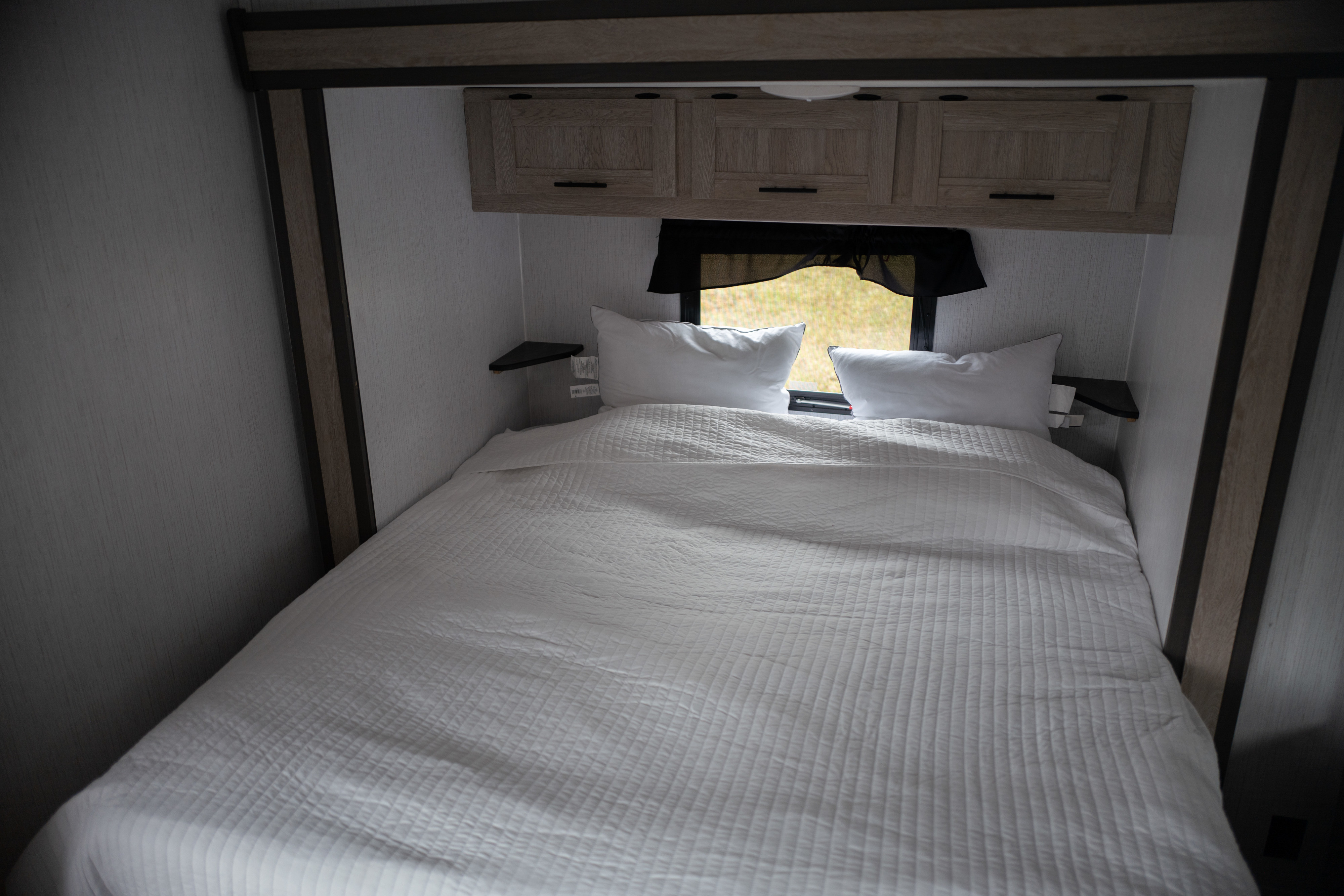 Bright RV rental bedroom with a comfy bed, white bedding, overhead storage, and a window view of nature.. Heartland Prowler 2022