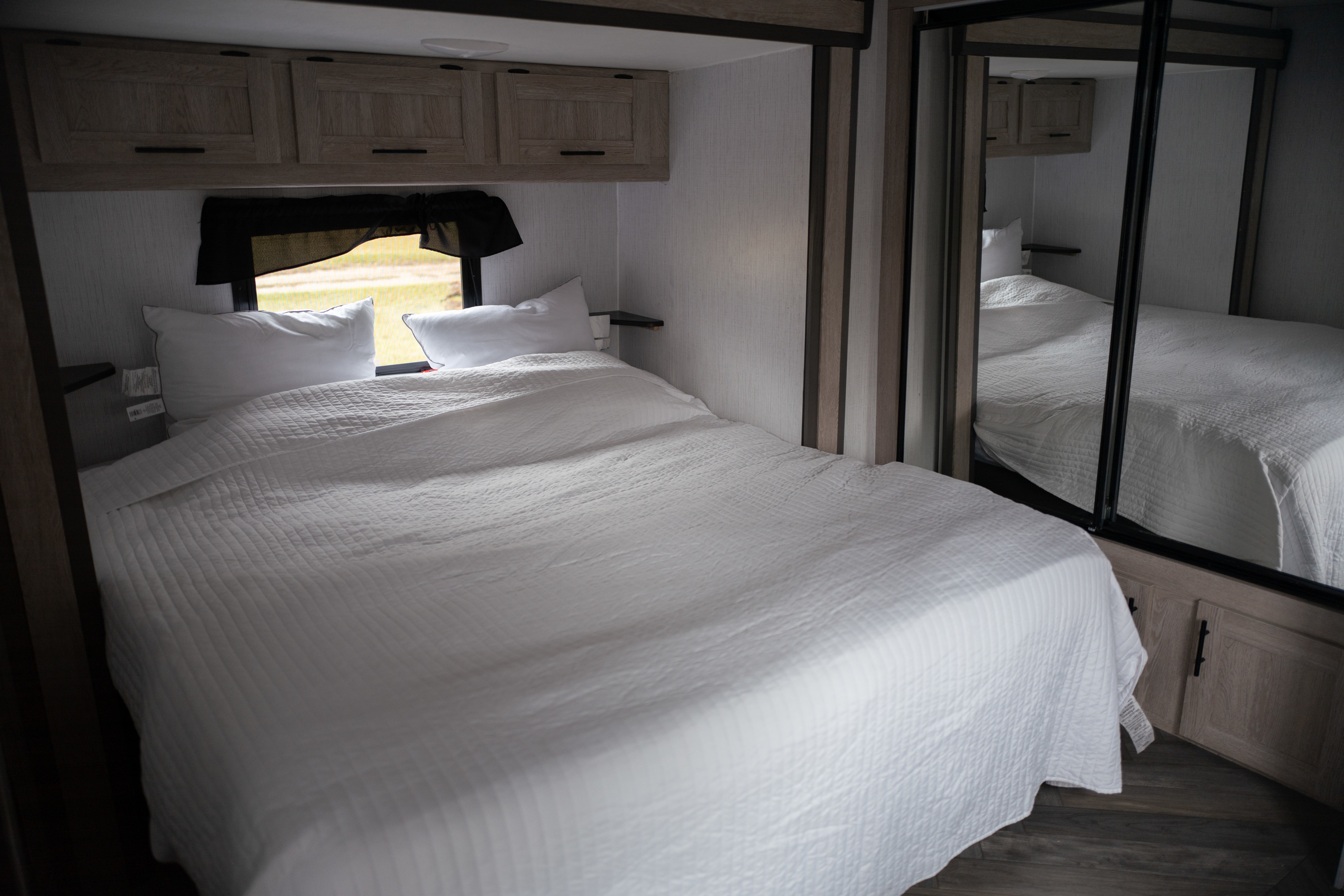Bright RV rental bedroom: large bed with white quilt, overhead cabinets, side shelves, window, and mirrored closet reflecting the room.. Heartland Prowler 2022