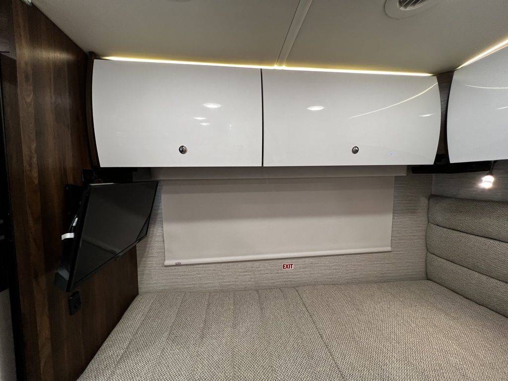 RV rental interior: modern bedroom with white overhead cabinets, wall-mounted TV, pull-down blind, and a patterned bed.. Winnebago View 2021