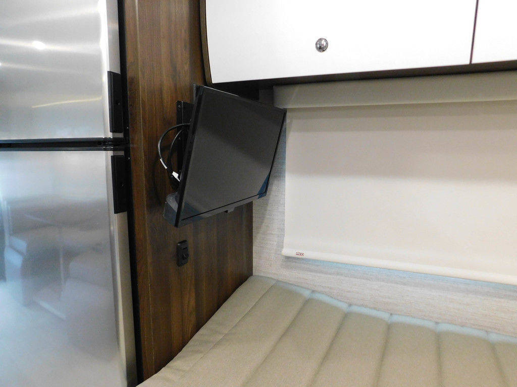 RV rental interior: stainless fridge, wall-mounted TV, white overhead cabinet, and a bed with window shade.. Winnebago View 2023