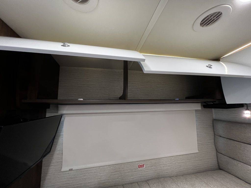 A bright RV rental interior with open white overhead cabinets, a TV, a pull-down exit shade, and cozy seating.. Winnebago View 2023