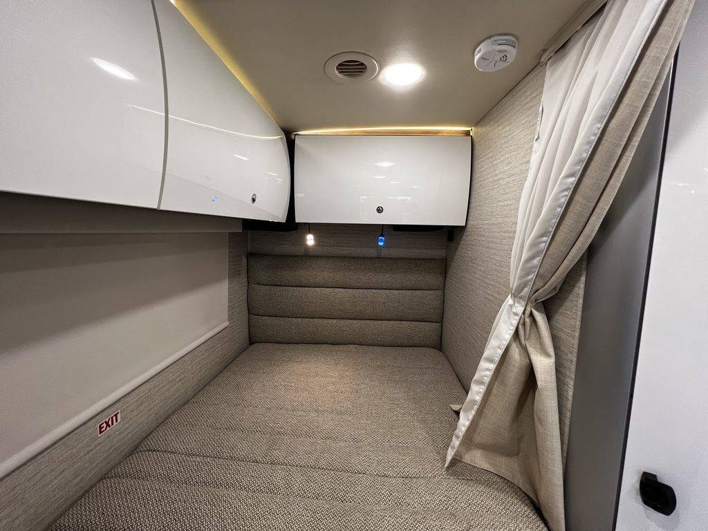 Inside a cozy RV rental: a compact sleeping area with modern overhead storage, an exit sign, and a tied-back curtain.. Winnebago View 2023