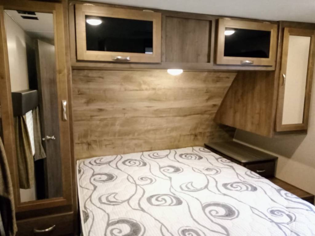 Cozy RV rental bedroom features a patterned bed, overhead cabinets, and a mirrored wardrobe for comfortable stays.. Dutchmen Coleman Light 2018