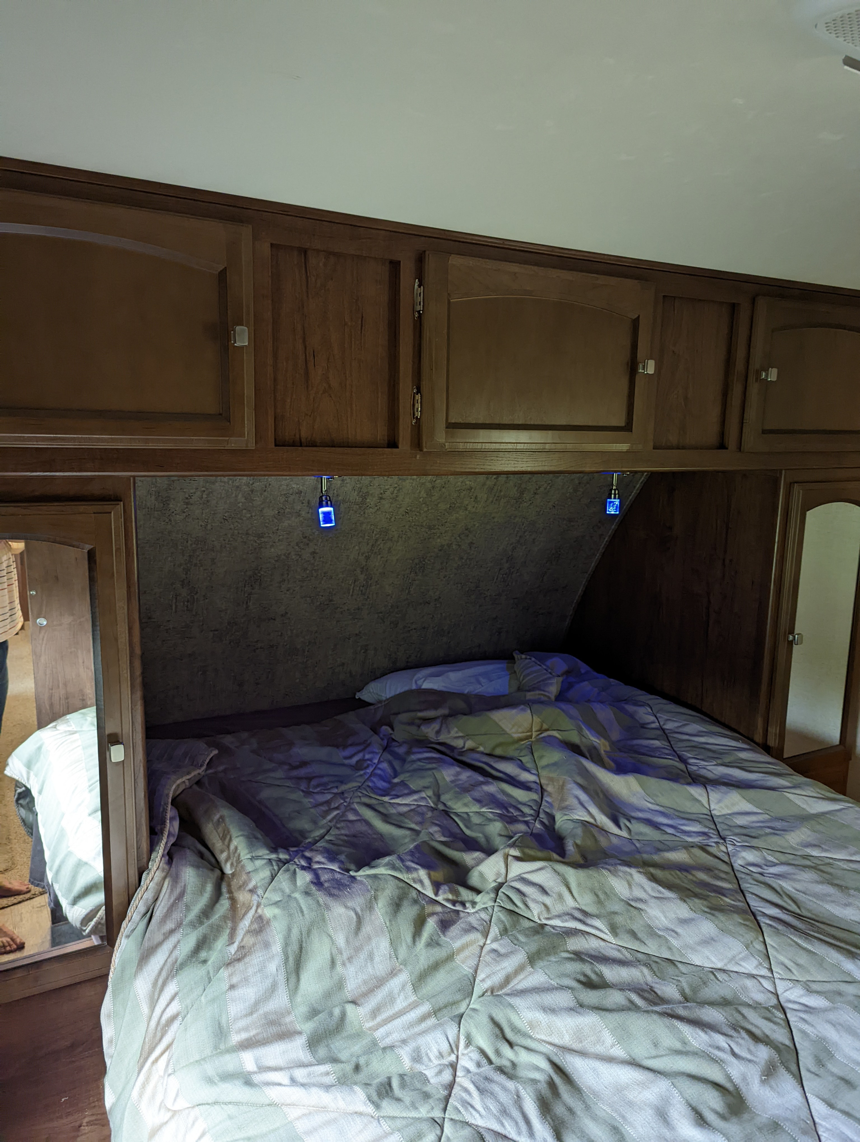 Interior of an RV rental bedroom showing a bed with a striped comforter, overhead wood cabinets, and glowing blue reading lights.. Forest River Coachmen Freedom Express 2017