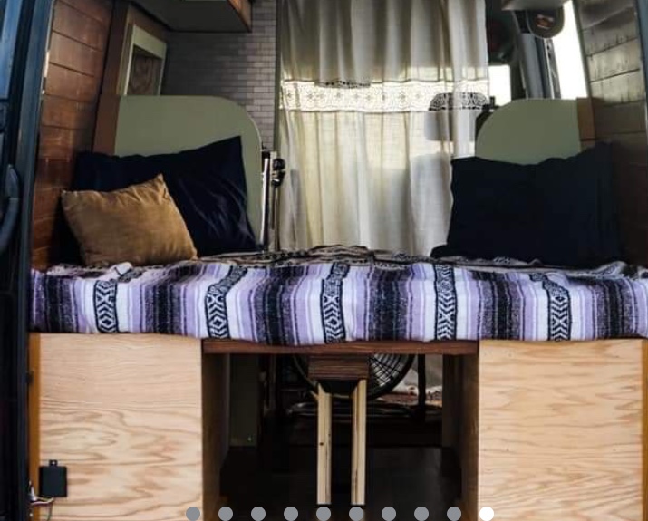 RV rental van interior: A cozy bed with a striped blanket & pillows, flanked by wood walls. Under-bed storage visible. White curtains in back.. Mercedes Sprinter 2013
