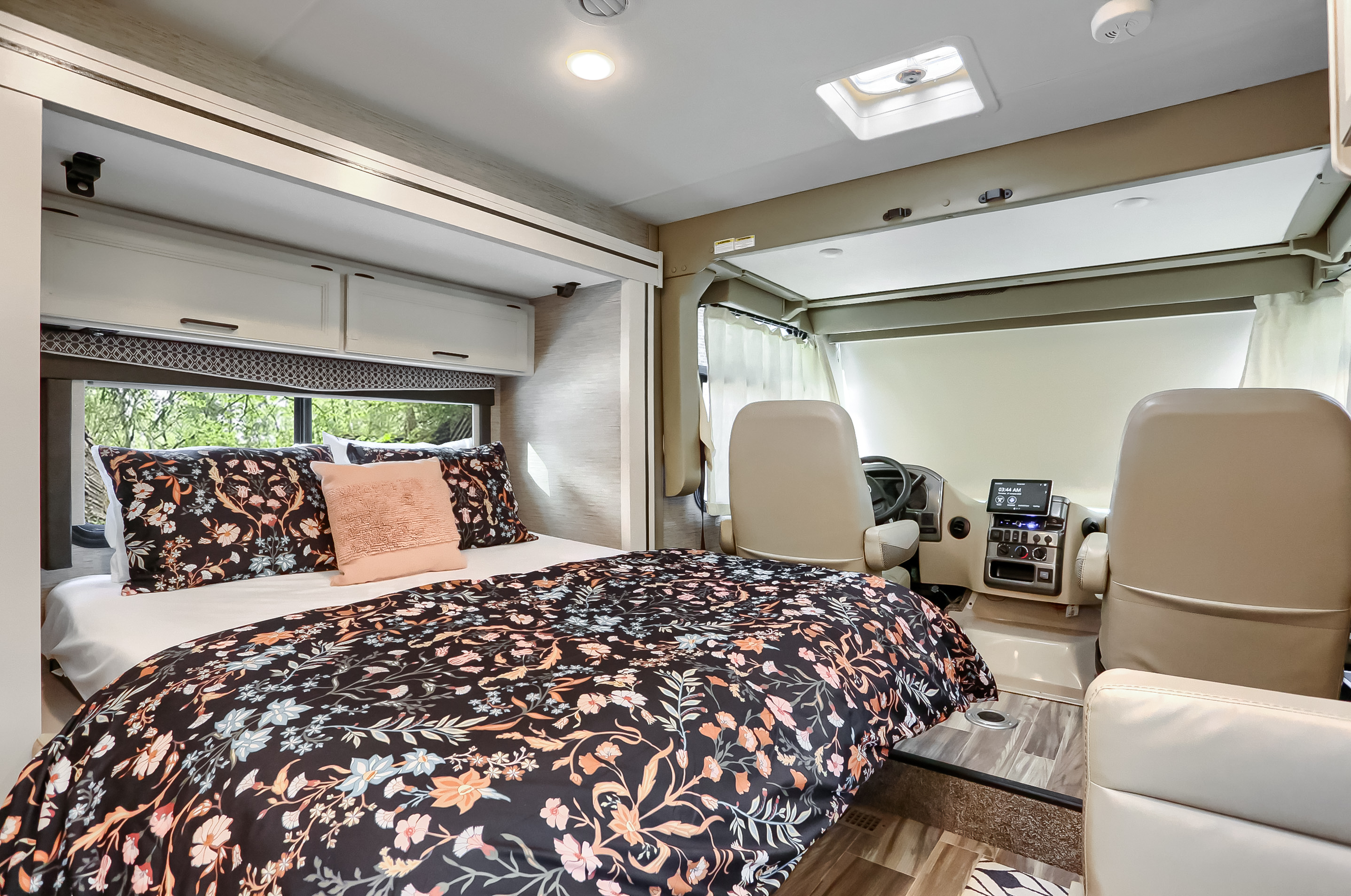 Inside an RV rental: a cozy bedroom with floral bedding seamlessly connects to the front cockpit area with tan seats.. Thor Vegas 2022