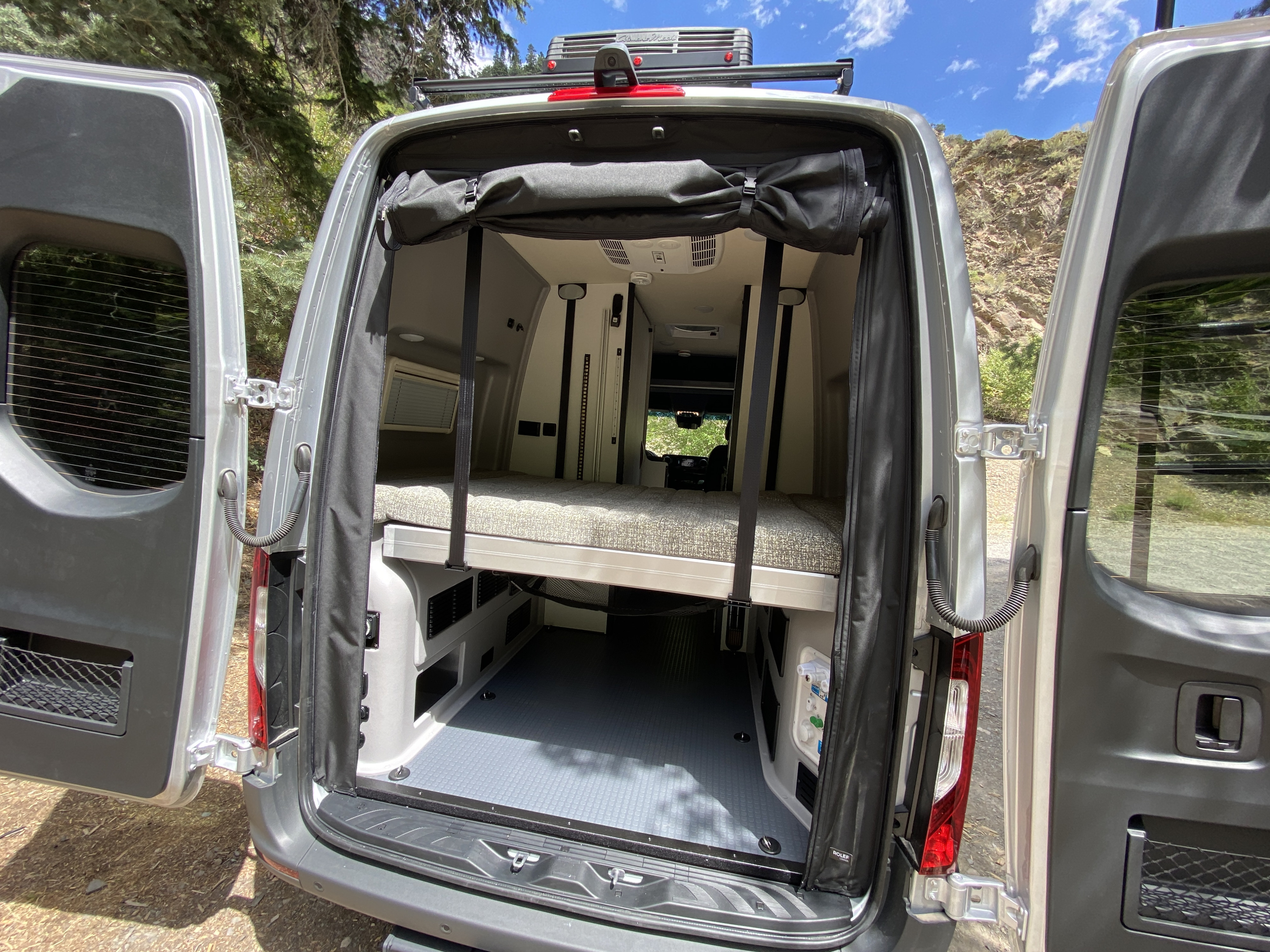 Discover this RV rental camper van's rear. Open doors reveal a raised bed, spacious storage below, and roll-up bug screens.. Winnebago Revel 2022