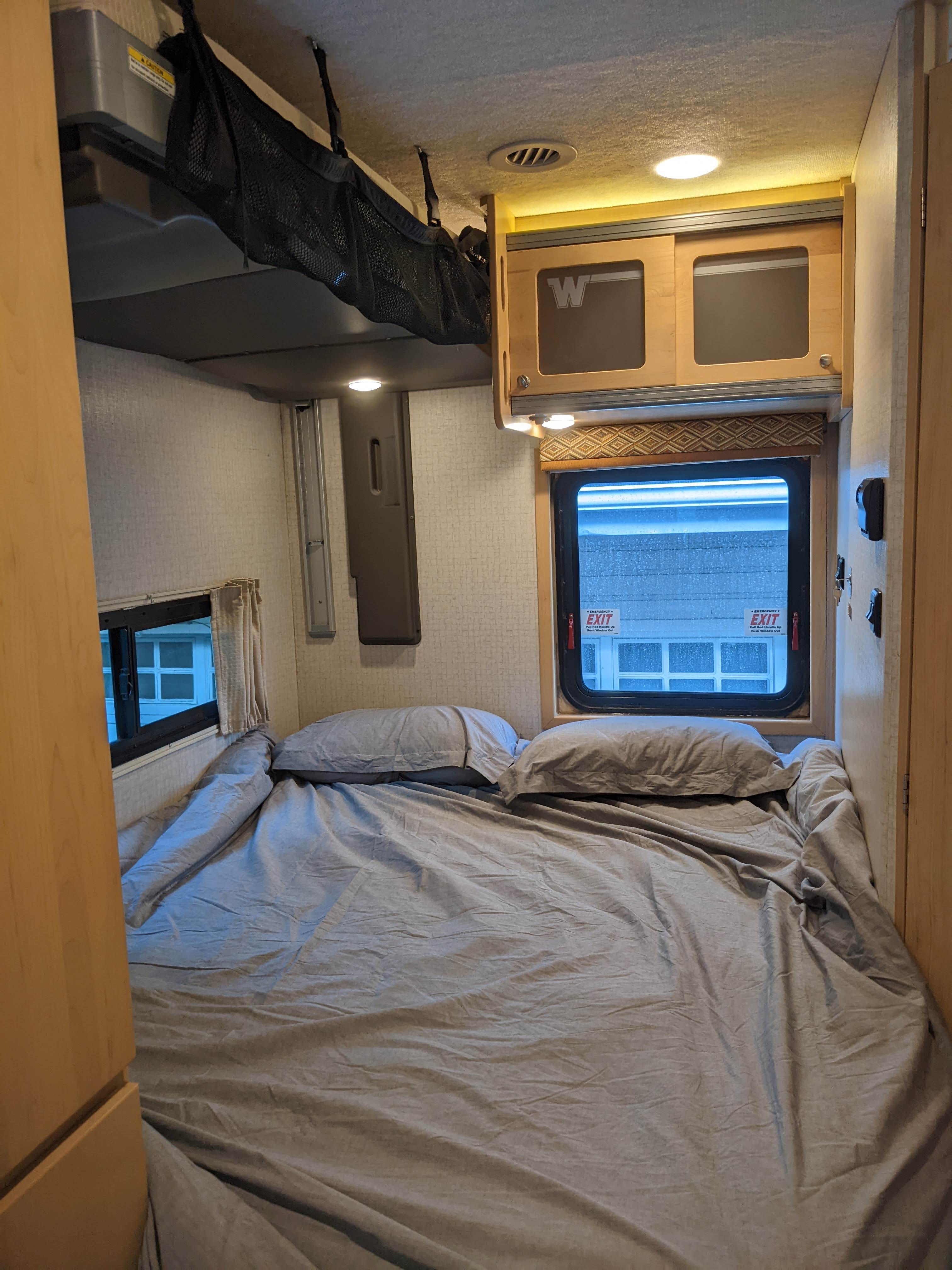 Inside an rv rental, a compact bedroom with a bed, grey linens, cabinets, and emergency exit windows.. Winnebago Brave 2015