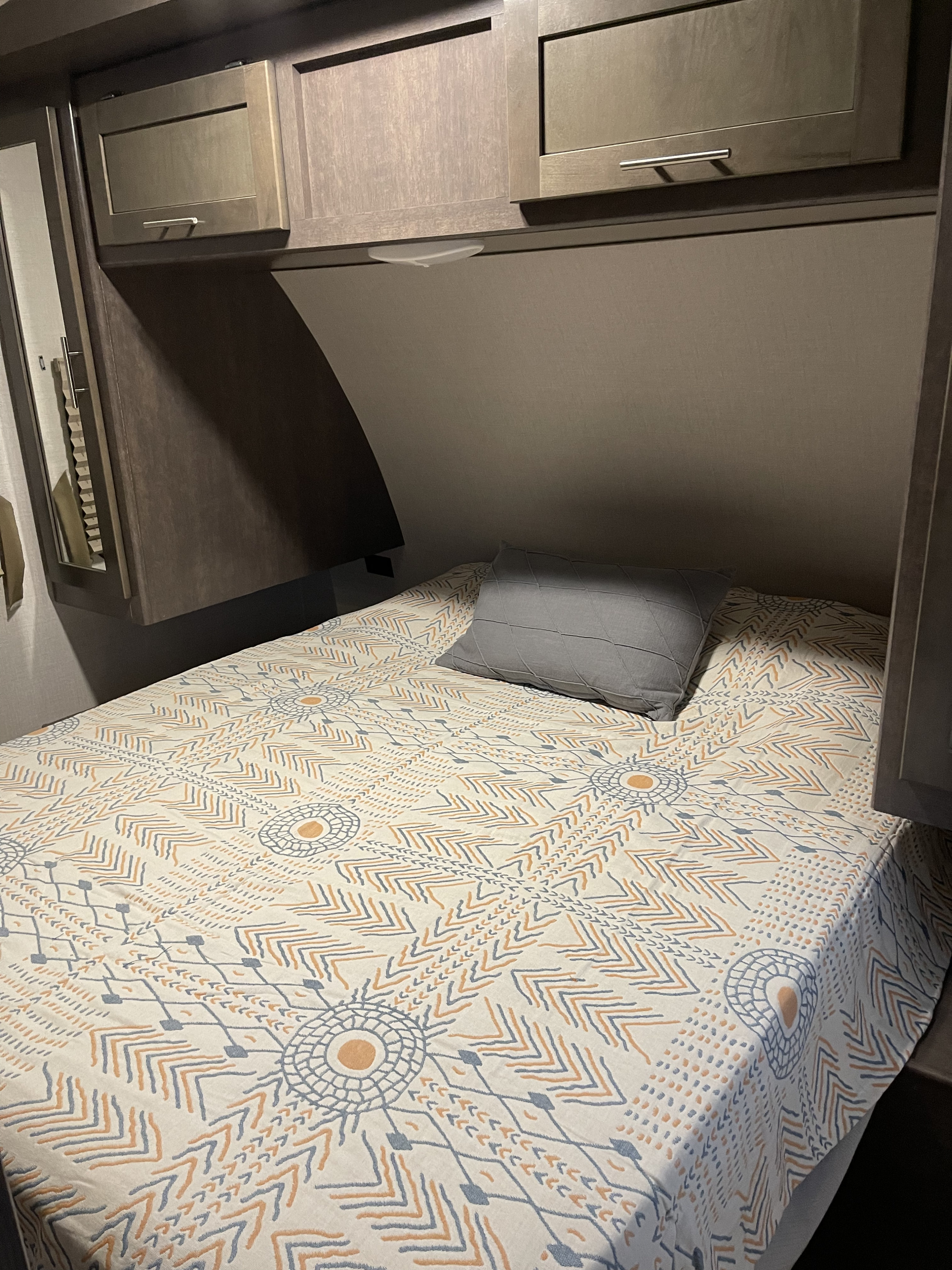 An RV rental's compact bedroom showing a bed with a white, orange, and grey patterned comforter, grey pillow, and wooden overhead cabinets.. Crossroads RV Sunset Trail Super Lite 2018