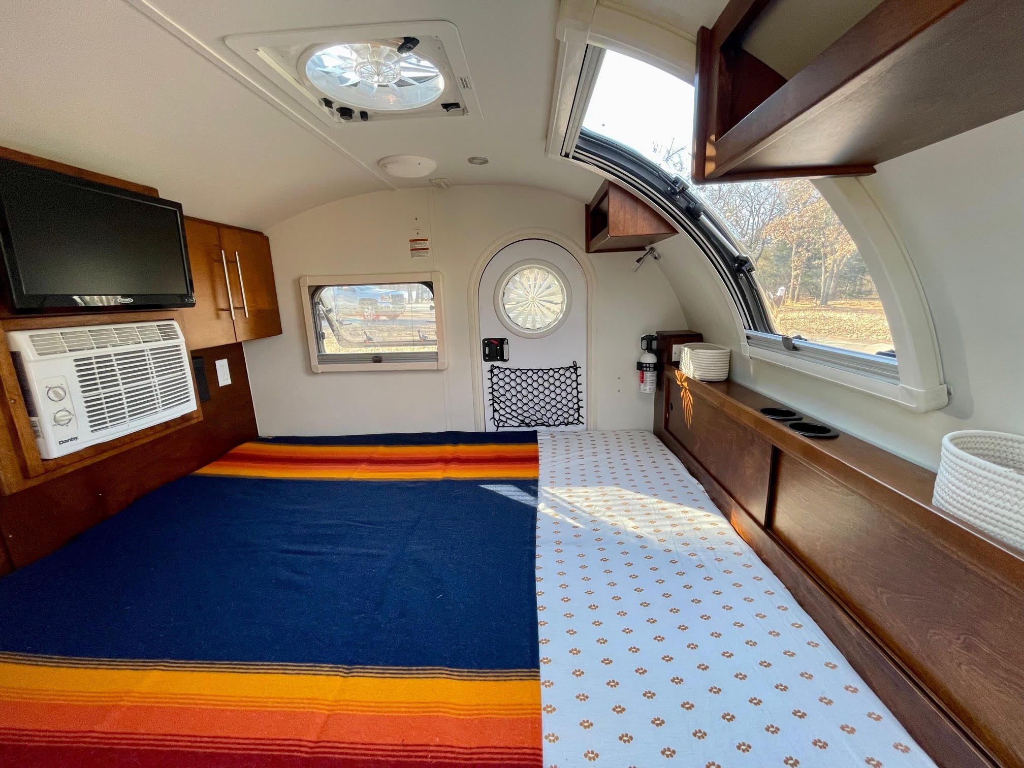 Bright RV rental interior displays a comfortable bed, TV, AC unit, and sunlit windows with outdoor scenery.. nuCamp T@G 2019
