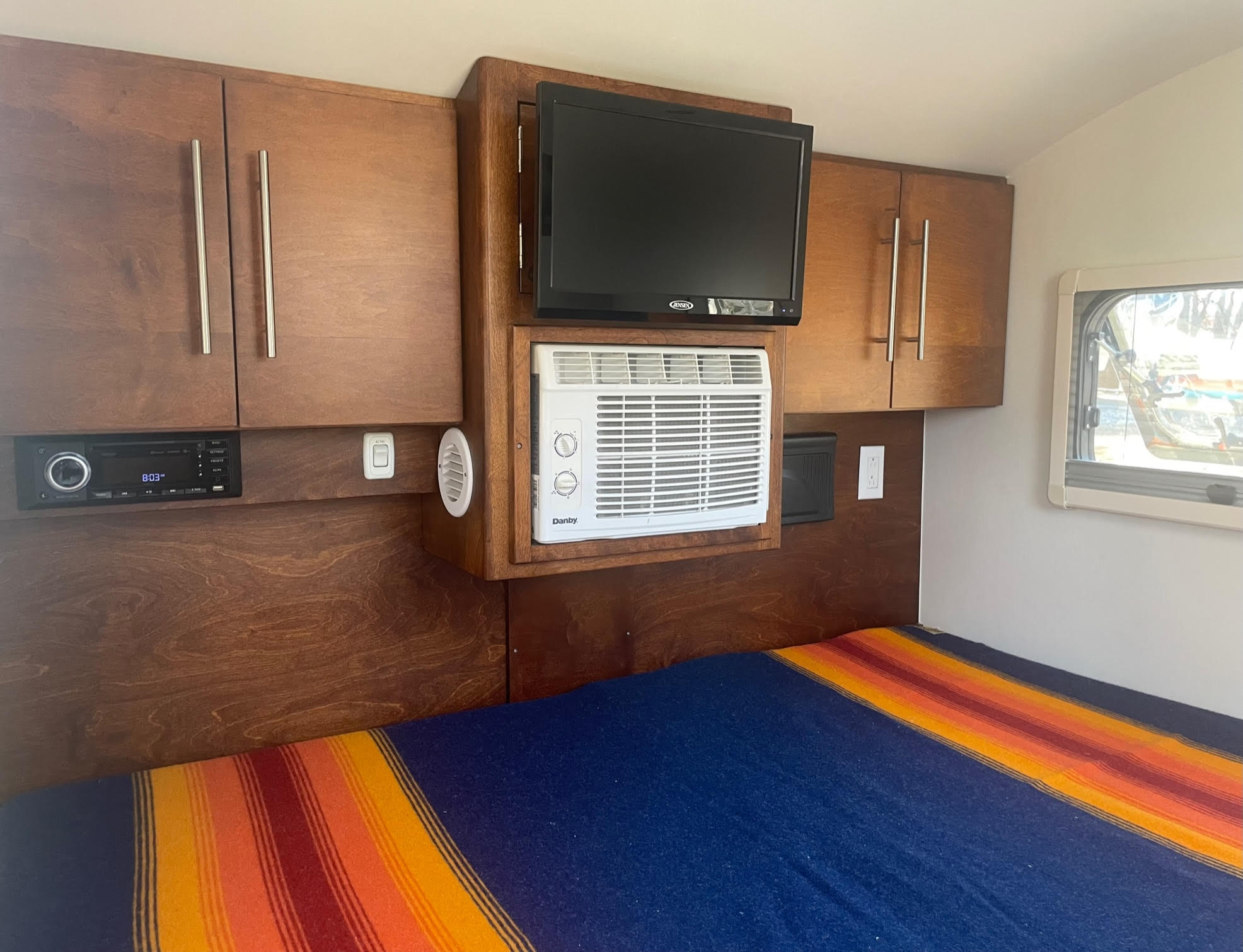 Experience comfort in this RV rental bedroom with warm wooden cabinets, an entertainment system, AC, and a vibrant bed setup.. nuCamp T@G 2019