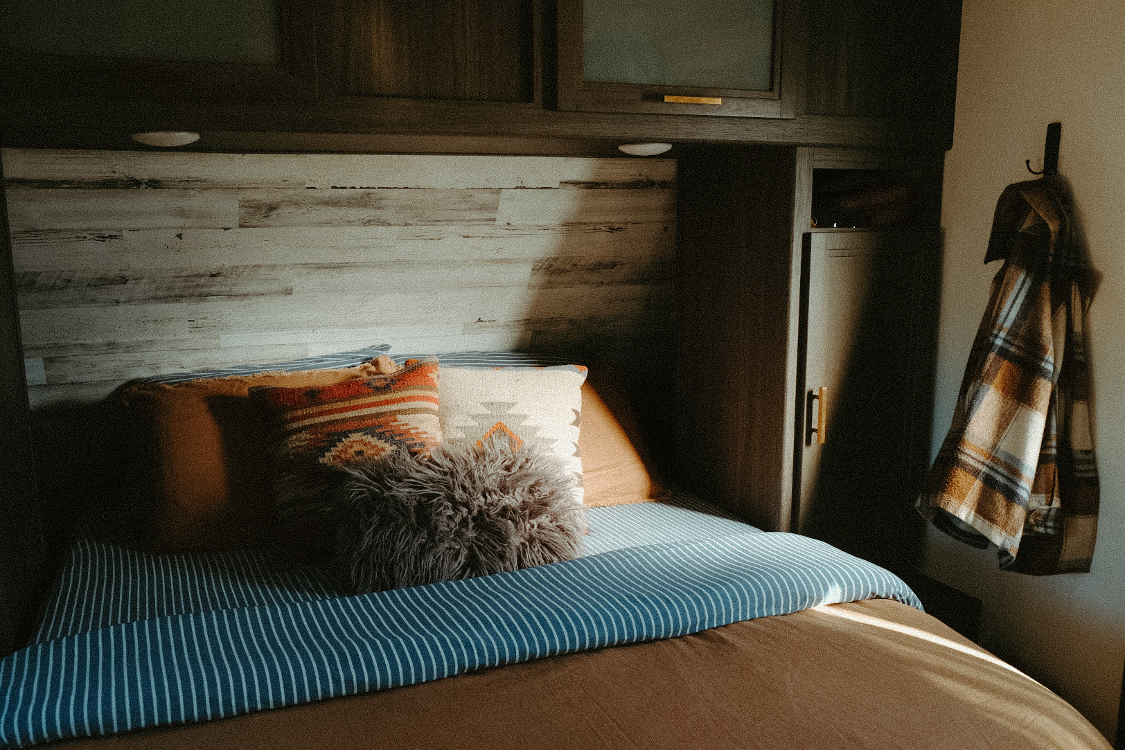 Cozy RV rental bedroom bathed in warm sunlight. Features a wood headboard, patterned pillows, and a hanging plaid shirt.. Dutchmen Kodiak Cub 2020