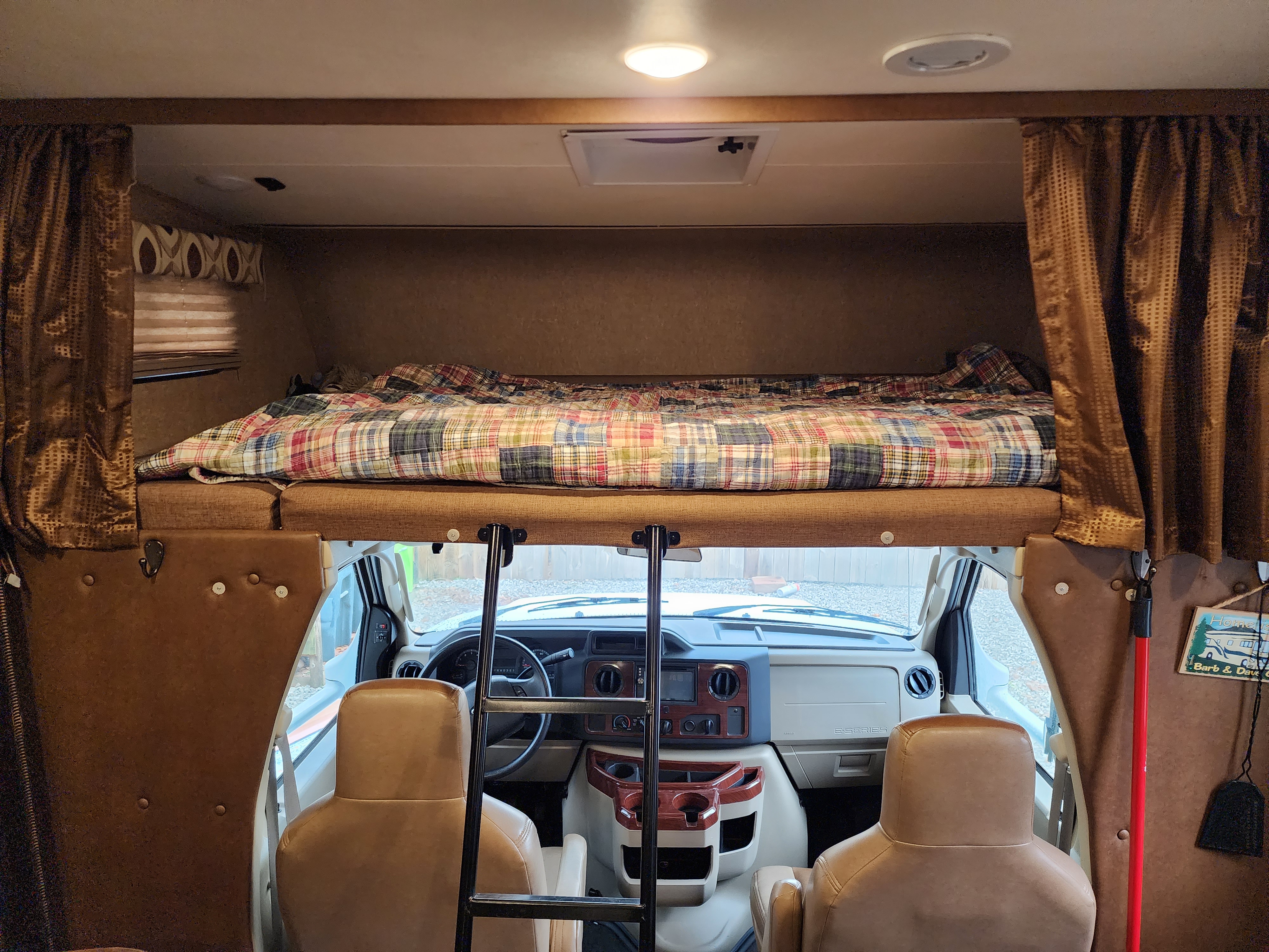 RV rental: Inside a motorhome, featuring a cozy cab-over bunk with plaid bedding, overlooking the driver's cockpit and seats.. Coach House Leprechaun 2015