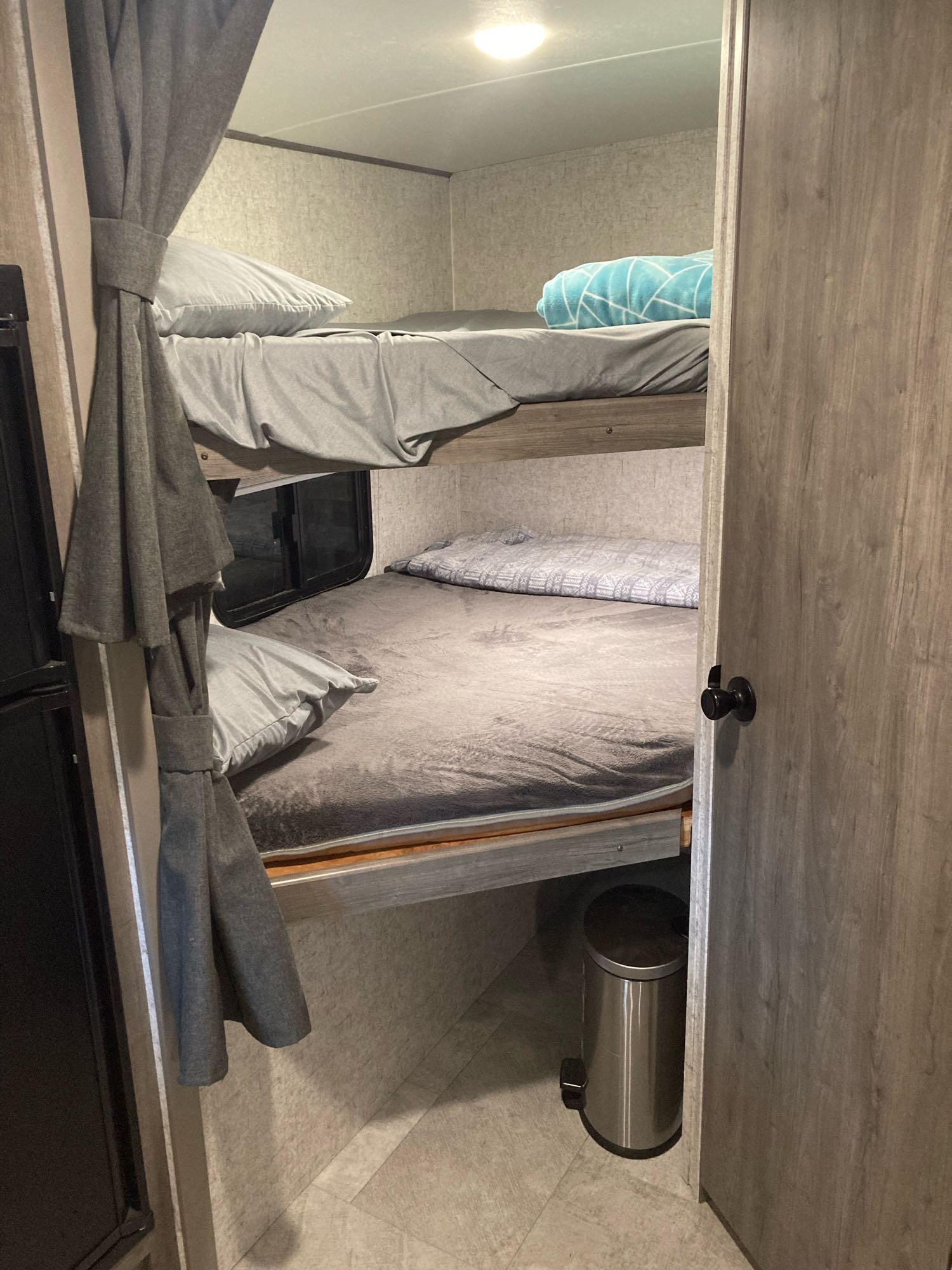 RV rental interior showing comfortable bunk beds. The top bunk has a blue blanket, while the bottom has a window, fridge, and small bin.. Coachmen Apex Nano 2020