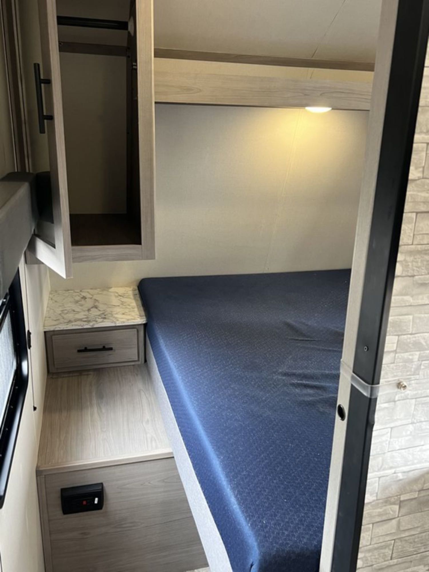 This RV rental interior shows a cozy sleeping area with a dark blue bed, nightstand, overhead cabinet, and reading light.. Dutchmen Coleman Lantern LT 2022