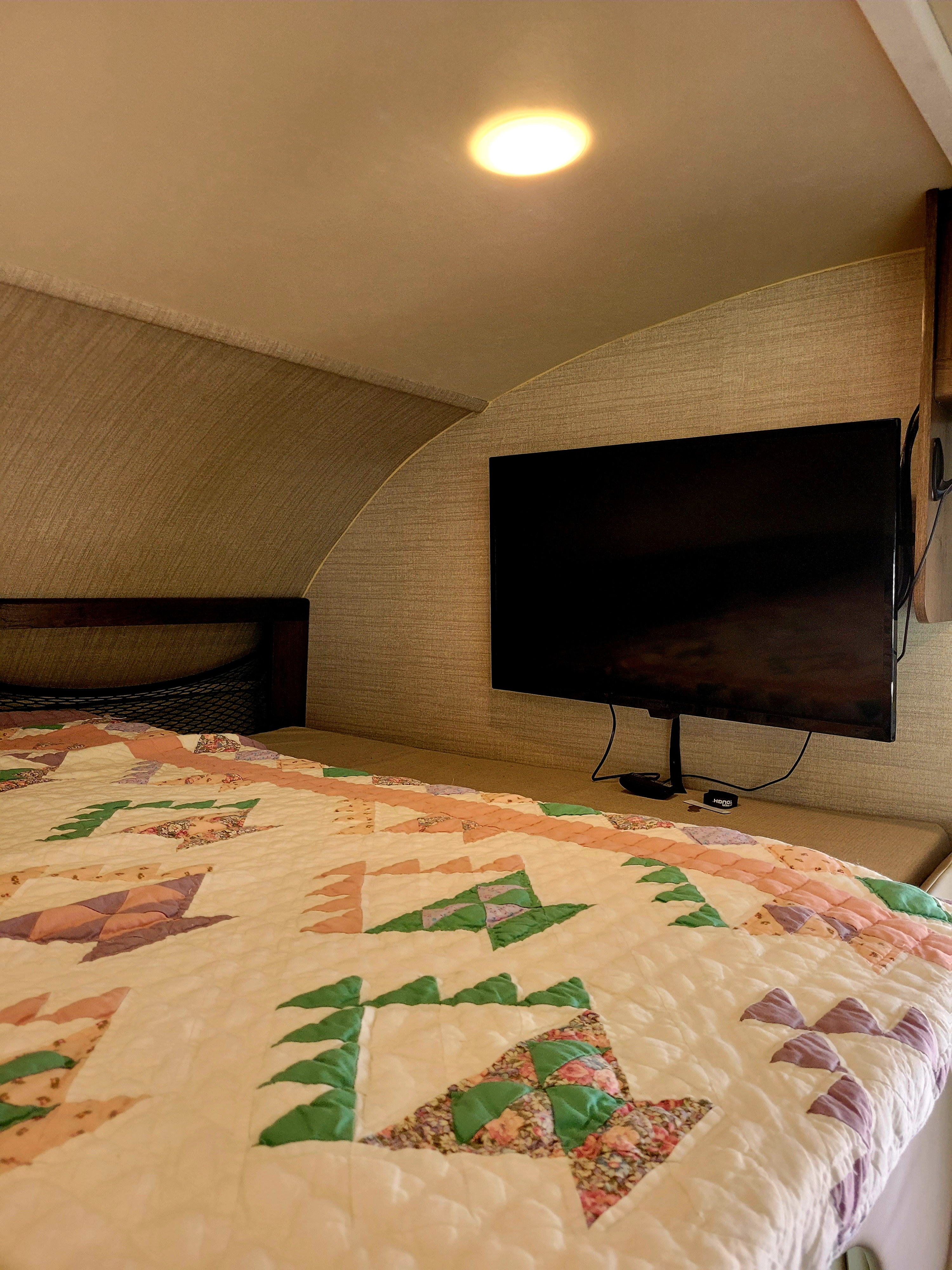 Inside a comfy RV rental: A sleeping area featuring a colorful quilt, wall-mounted TV, and overhead light.. Jayco Redhawk 2020