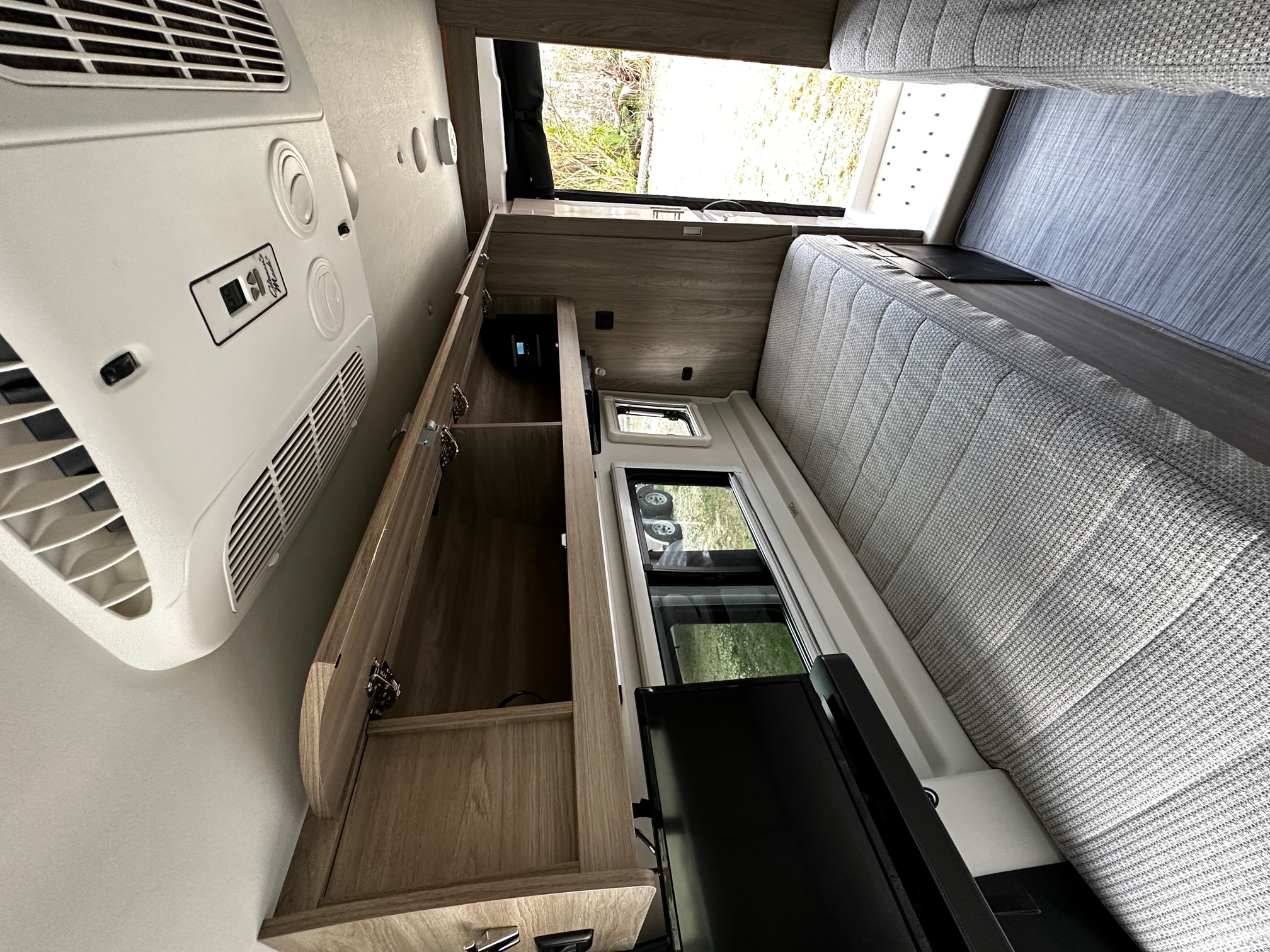 RV rental interior: Overhead AC unit, light wood cabinets, a long bed/seating area, and side windows providing natural light.. Winnebago Travato 2022