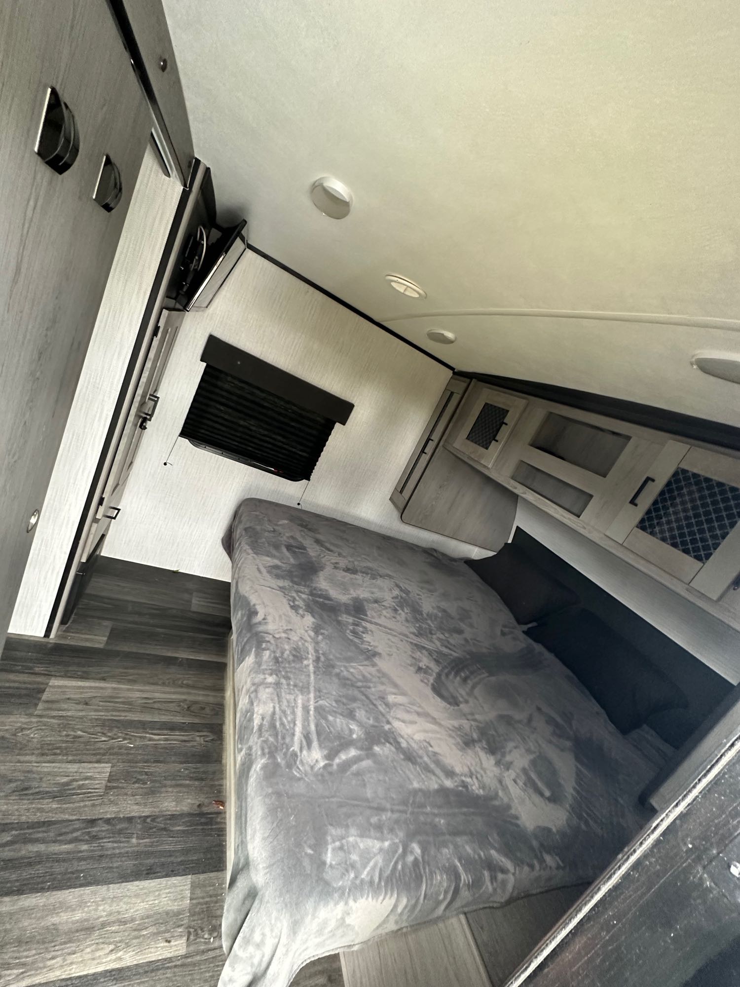 Inside an RV rental bedroom: gray bed with blanket, overhead cabinets, window, and dark wood-look floor.. Heartland Other 2021