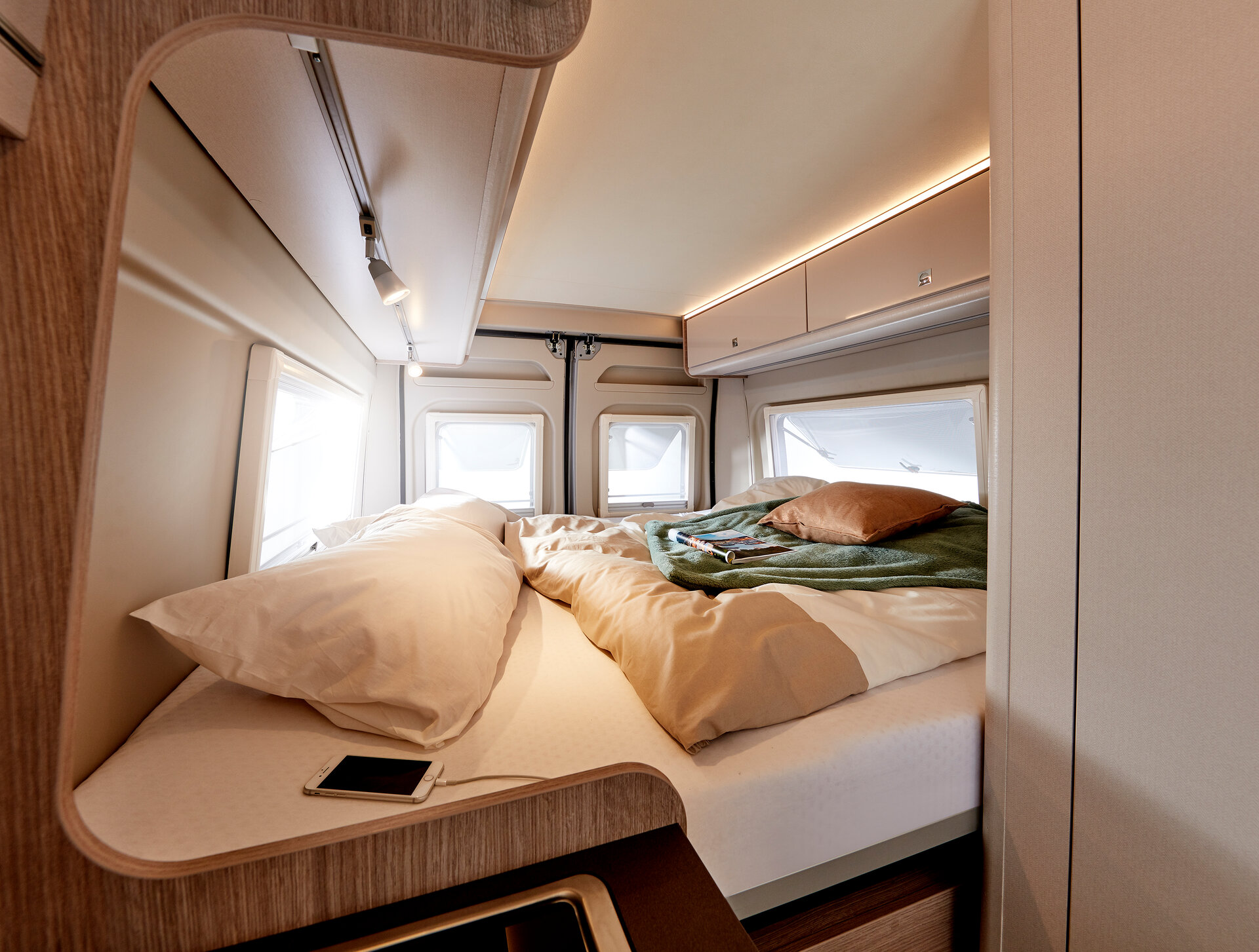 Spacious and modern RV rental bedroom features a comfy bed with bright windows, ambient lighting, and a smartphone charging.. Bürstner CAMPEO C600 2023