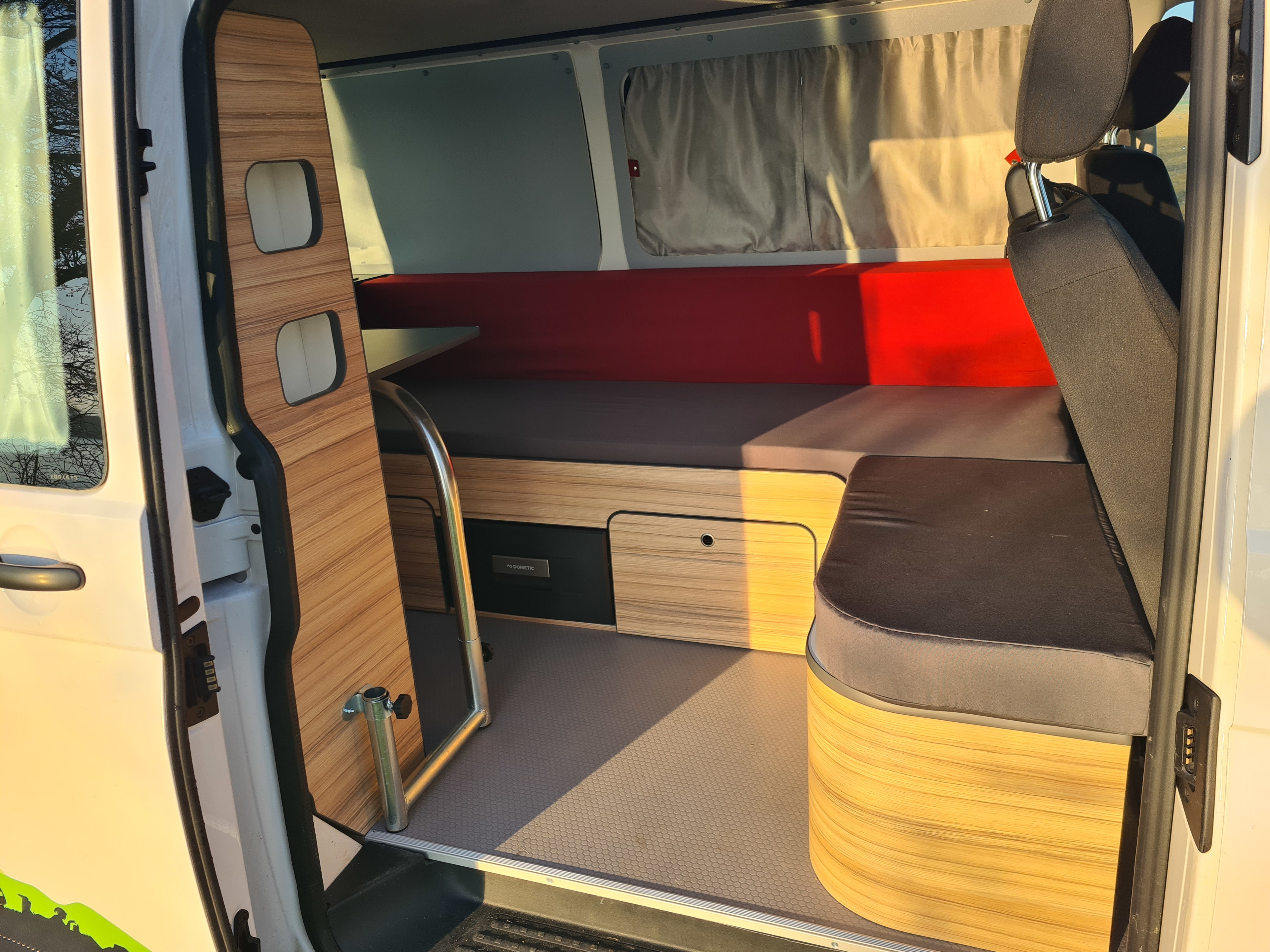 Bright RV rental interior featuring wooden cabinets, red and grey cushions for a bed/seating, and a pull-out table.. Volkswagen Transporter 2021