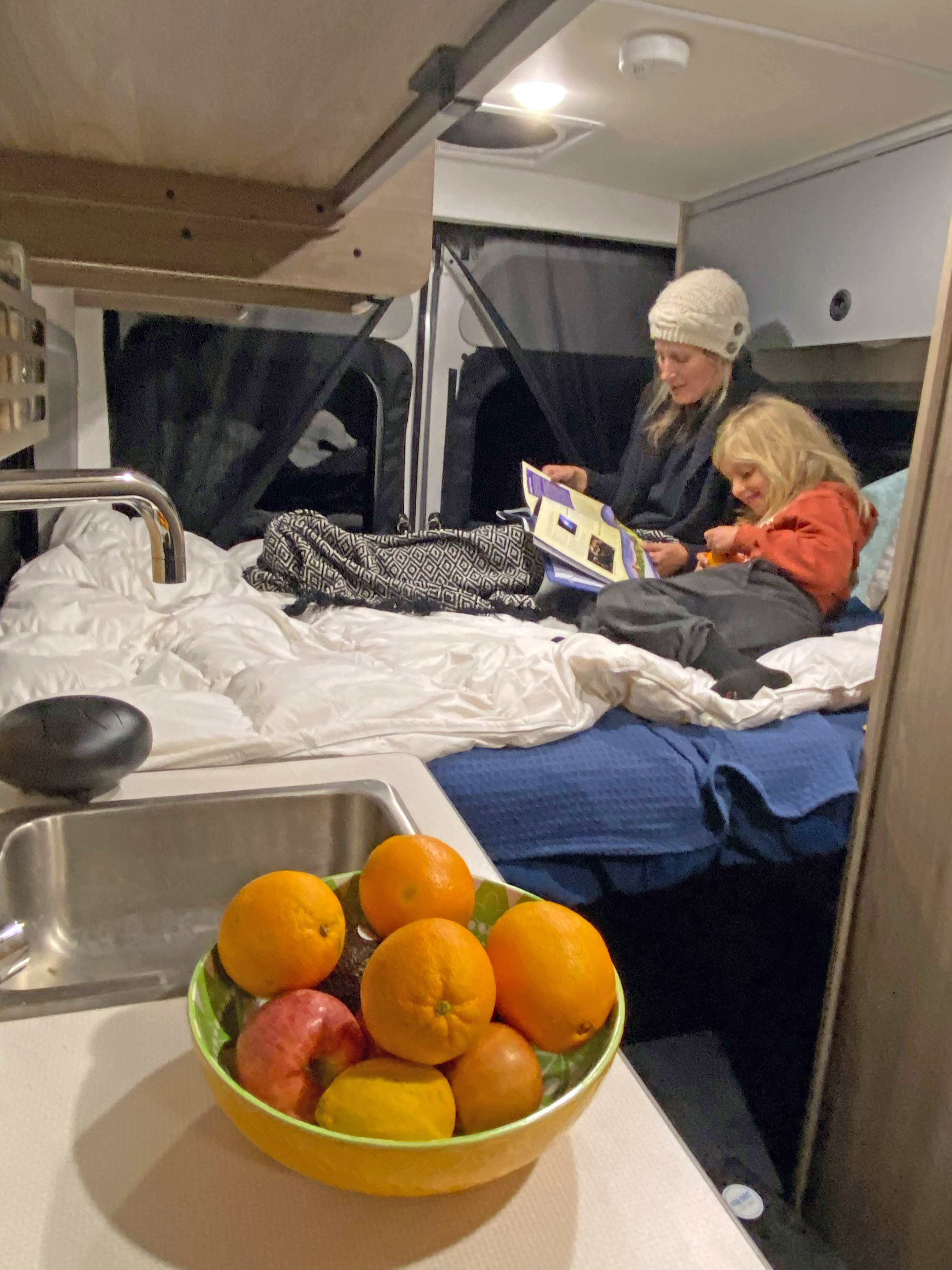 Cozy RV rental: Mother and child reading on the bed. A bowl of fresh fruit is in the foreground.. Winnebago Solis 2023