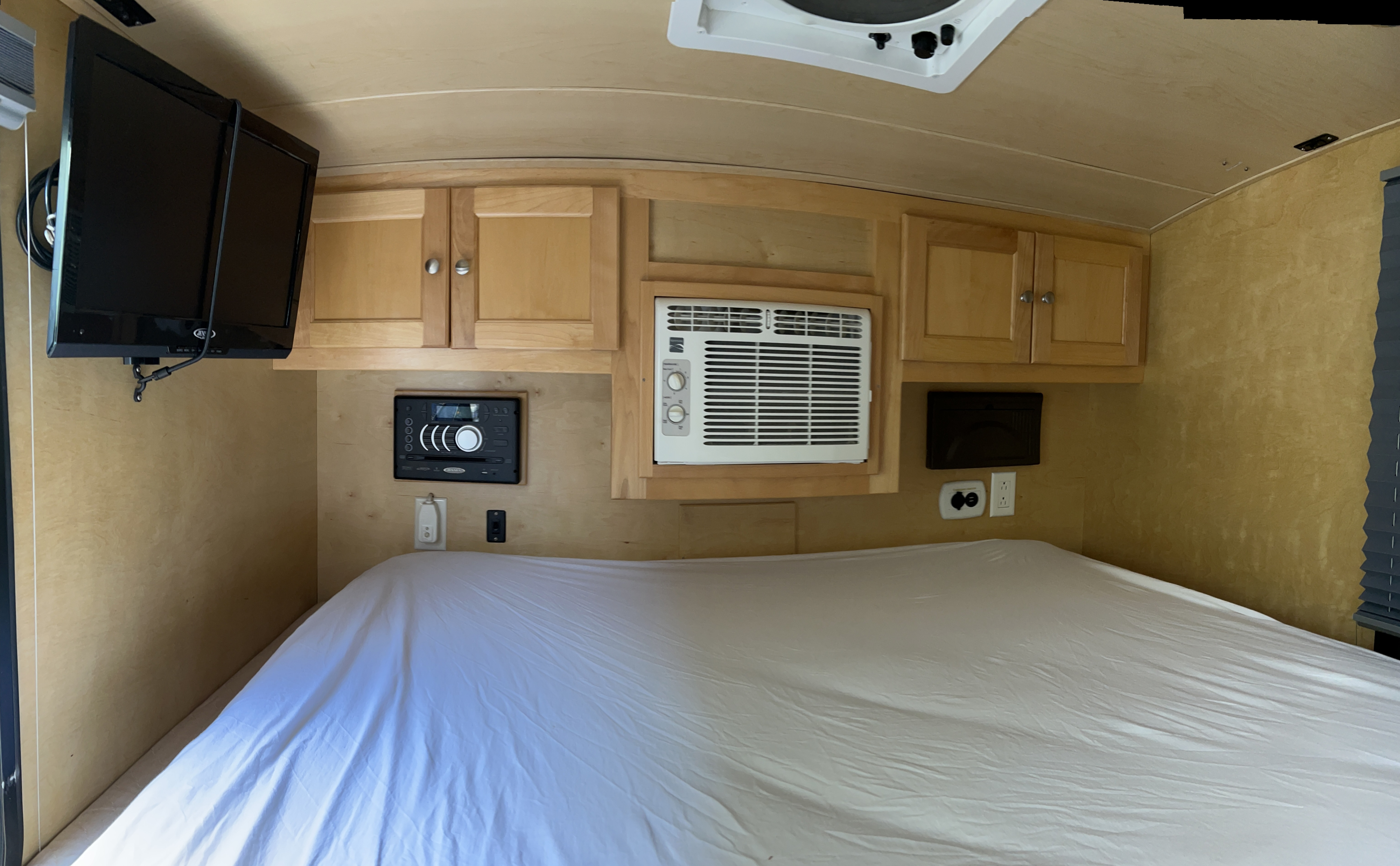 This cozy RV rental interior features a bed with white sheets, wall-mounted TV, stereo, AC unit, and wooden storage cabinets.. Little Guy Silver Shadow 2016