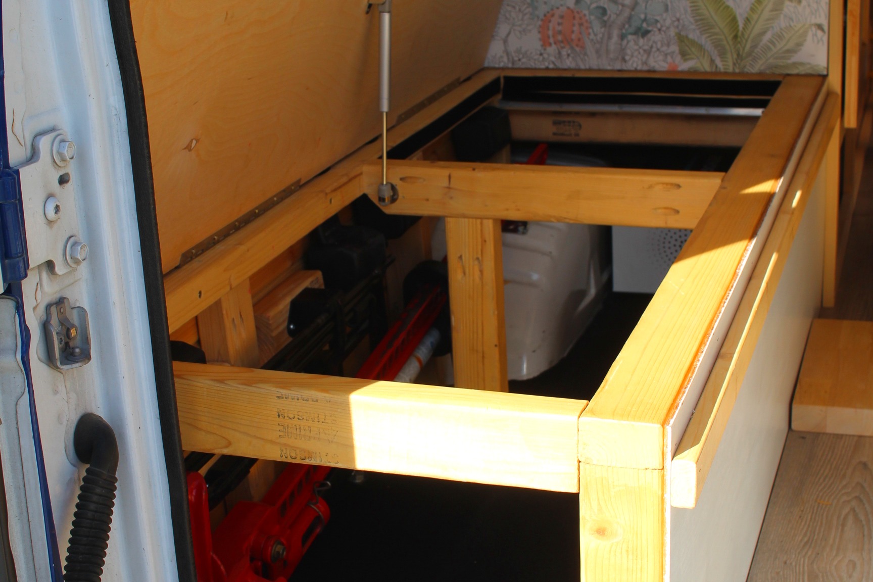 Inside an RV rental: a wooden bed frame with a hinged panel supported by a gas strut, revealing practical under-bed storage.. Ford Ford E350 2003