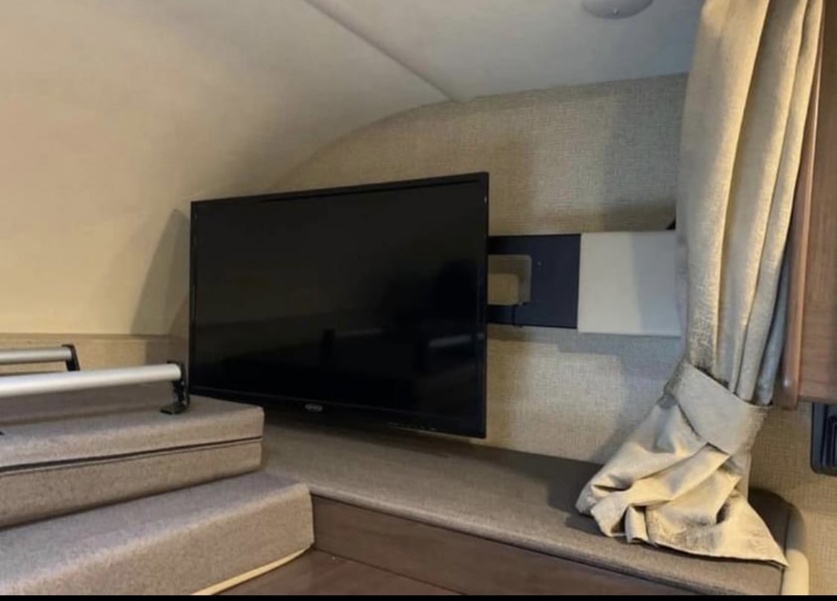 Spacious RV rental interior with a wall-mounted TV over carpeted steps leading to an elevated bunk. A curtain is on right.. Thor Four Winds 2019