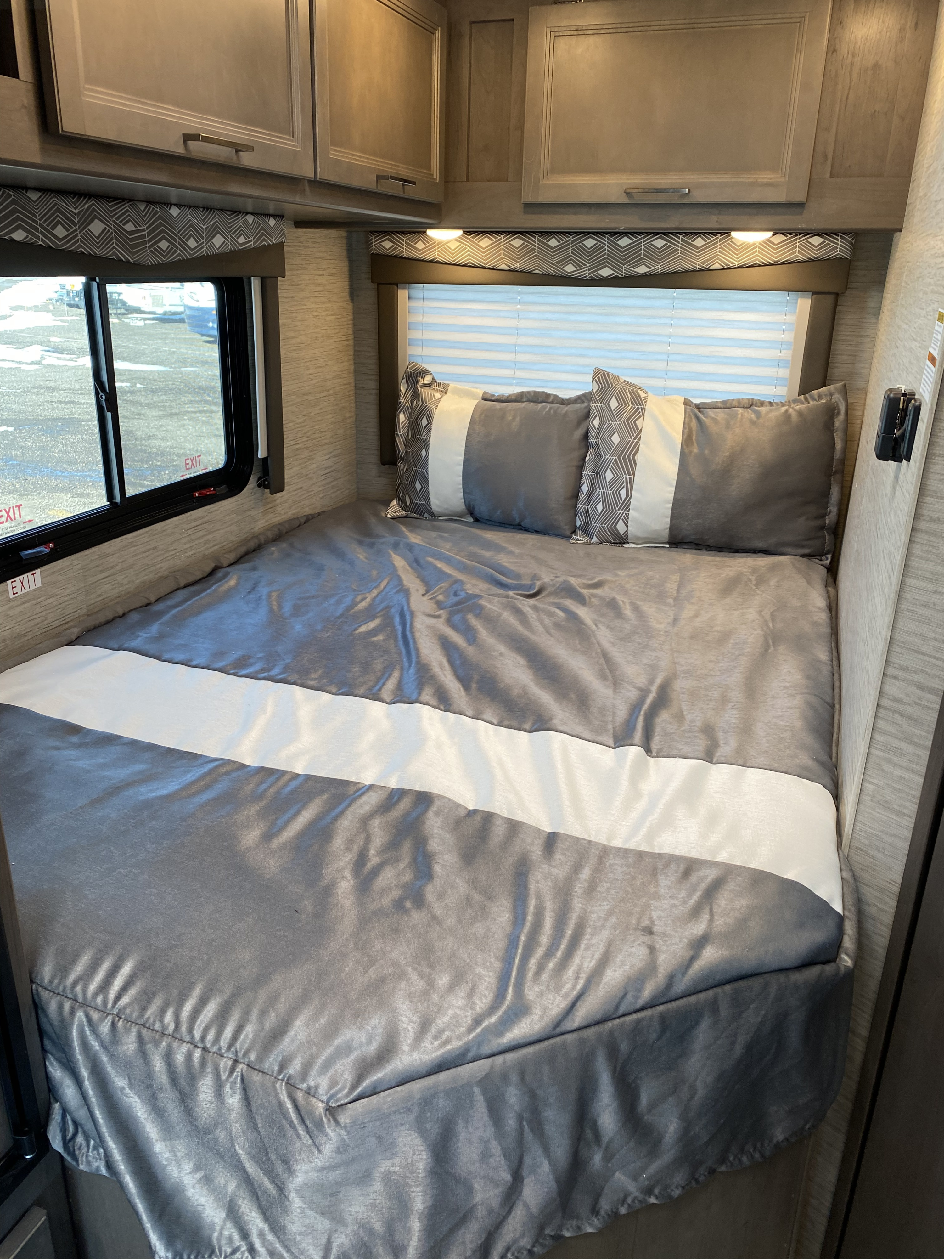 Cozy RV rental bedroom featuring a neatly made bed with grey and white bedding, decorative pillows, and overhead cabinets.. Thor Motor Coach Four Winds 2024