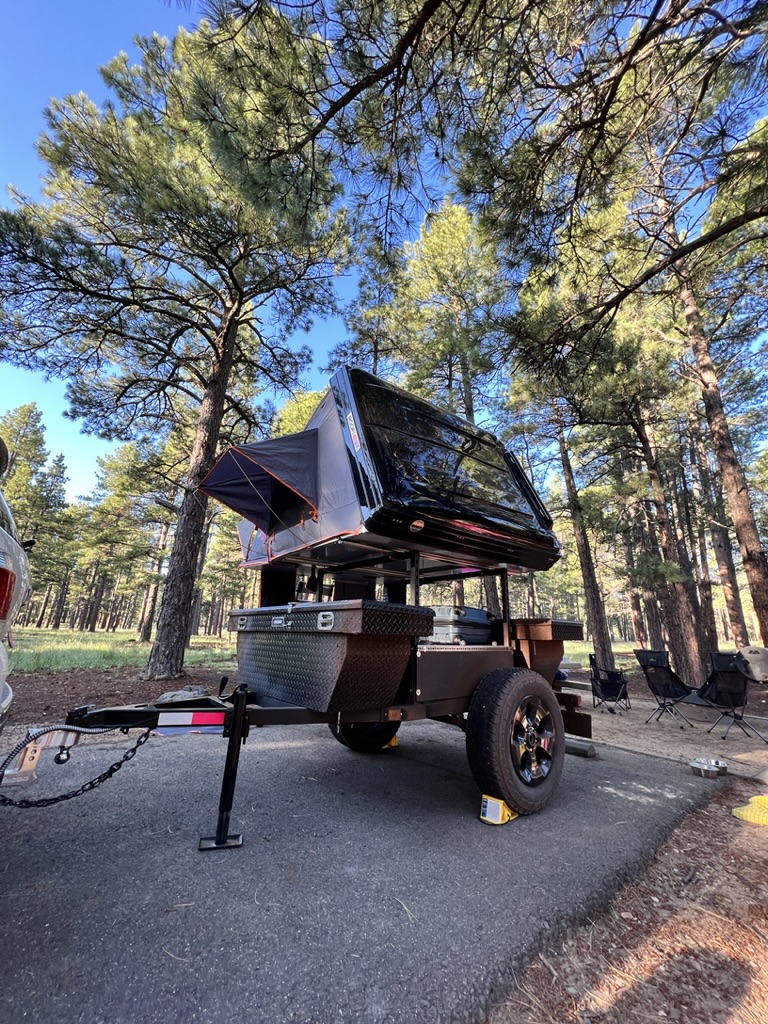A dark overland trailer with a deployed rooftop tent, an RV rental alternative, parked at a forest campsite.. Ady ORT 2022