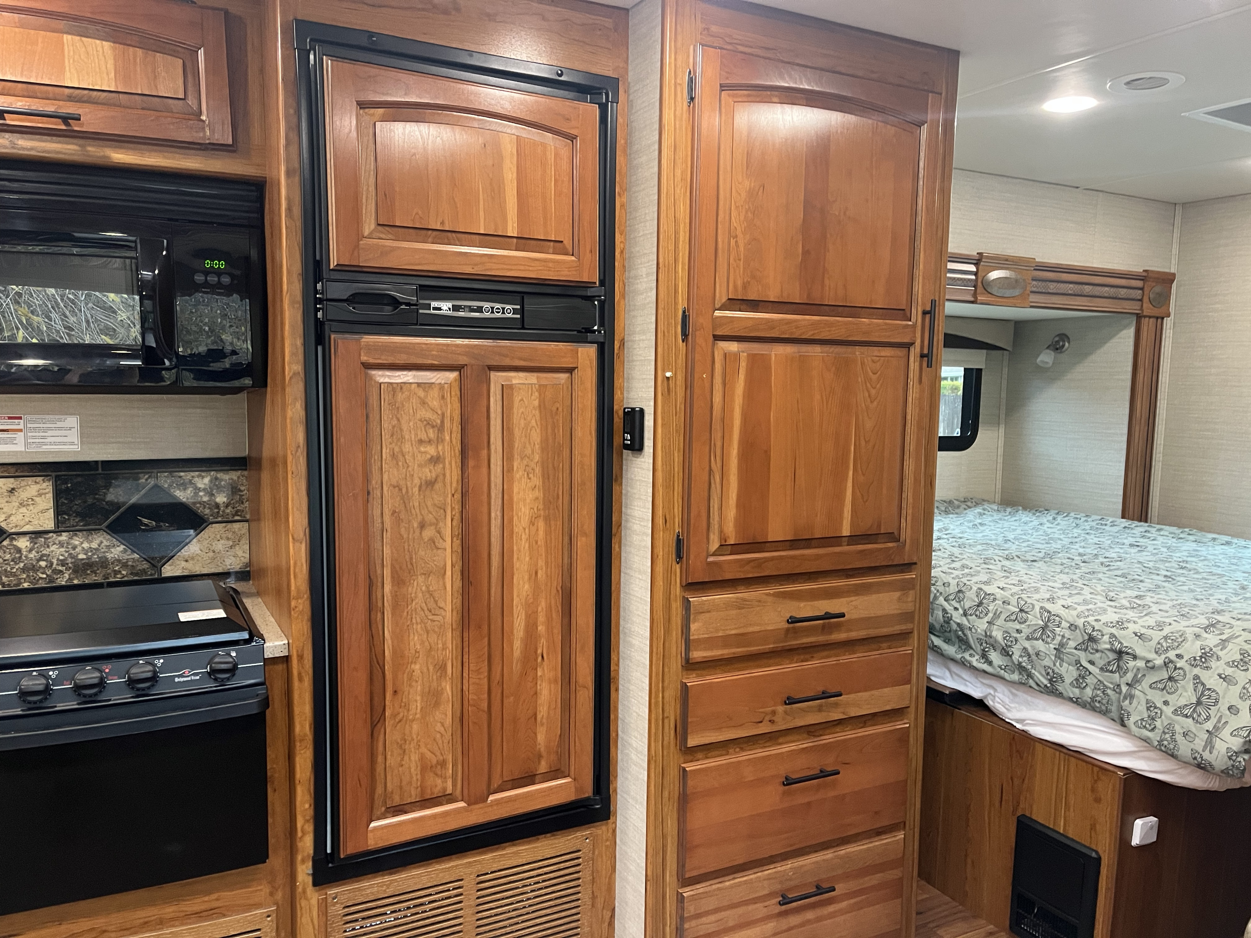 RV rental interior: A compact kitchen with microwave, fridge, and stove, next to a cozy bedroom with patterned bedding and storage.. Jayco Greyhawk 2017