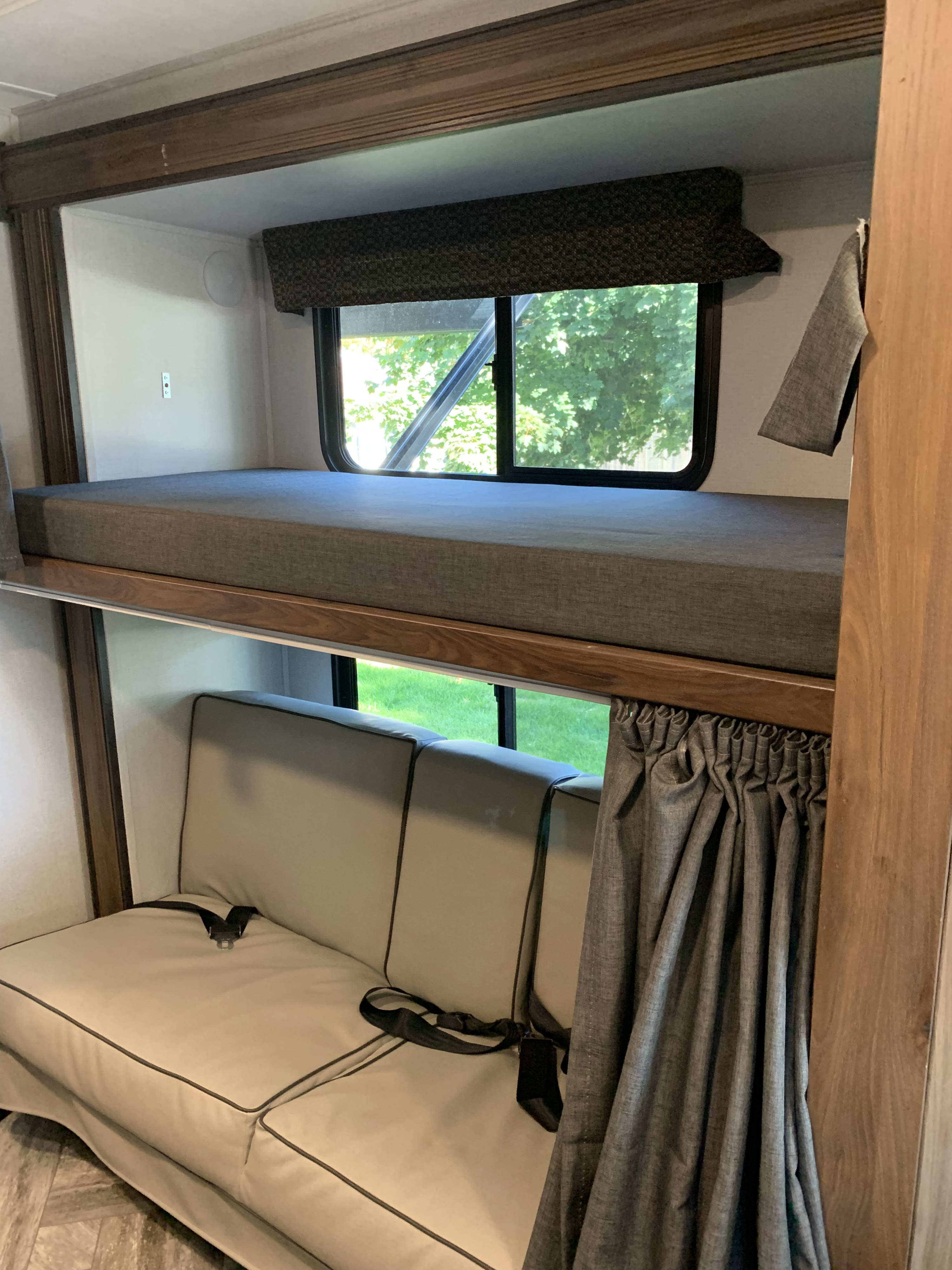 RV rental interior: a bunk bed with gray mattress above a beige sofa with seatbelts, surrounded by wood trim and windows.. Forest River Sunseeker LE 2022