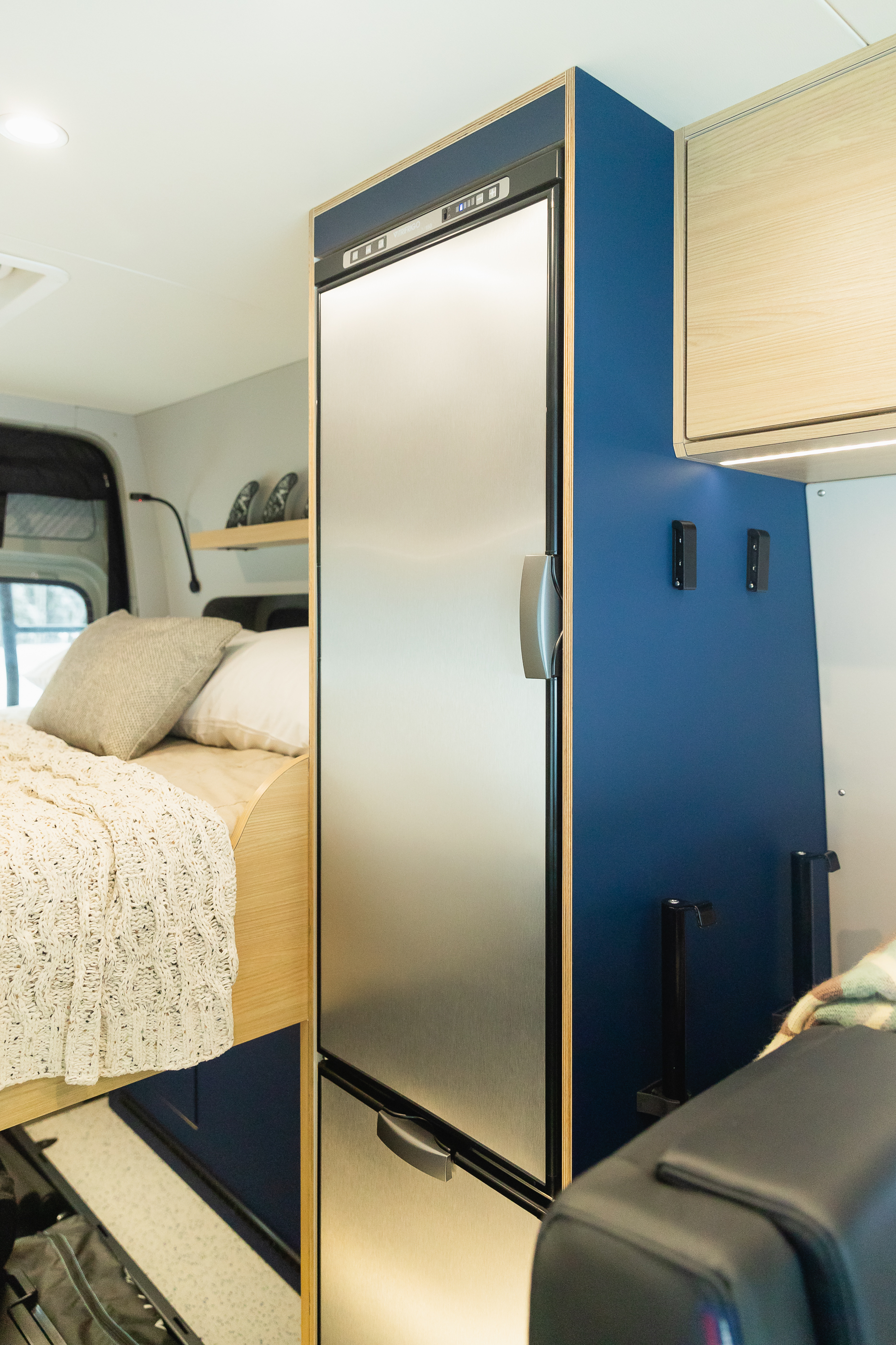 This RV rental features a tall stainless steel fridge integrated into blue and wood custom cabinetry, next to a cozy bed.. Mercedes-Benz Sprinter 2022