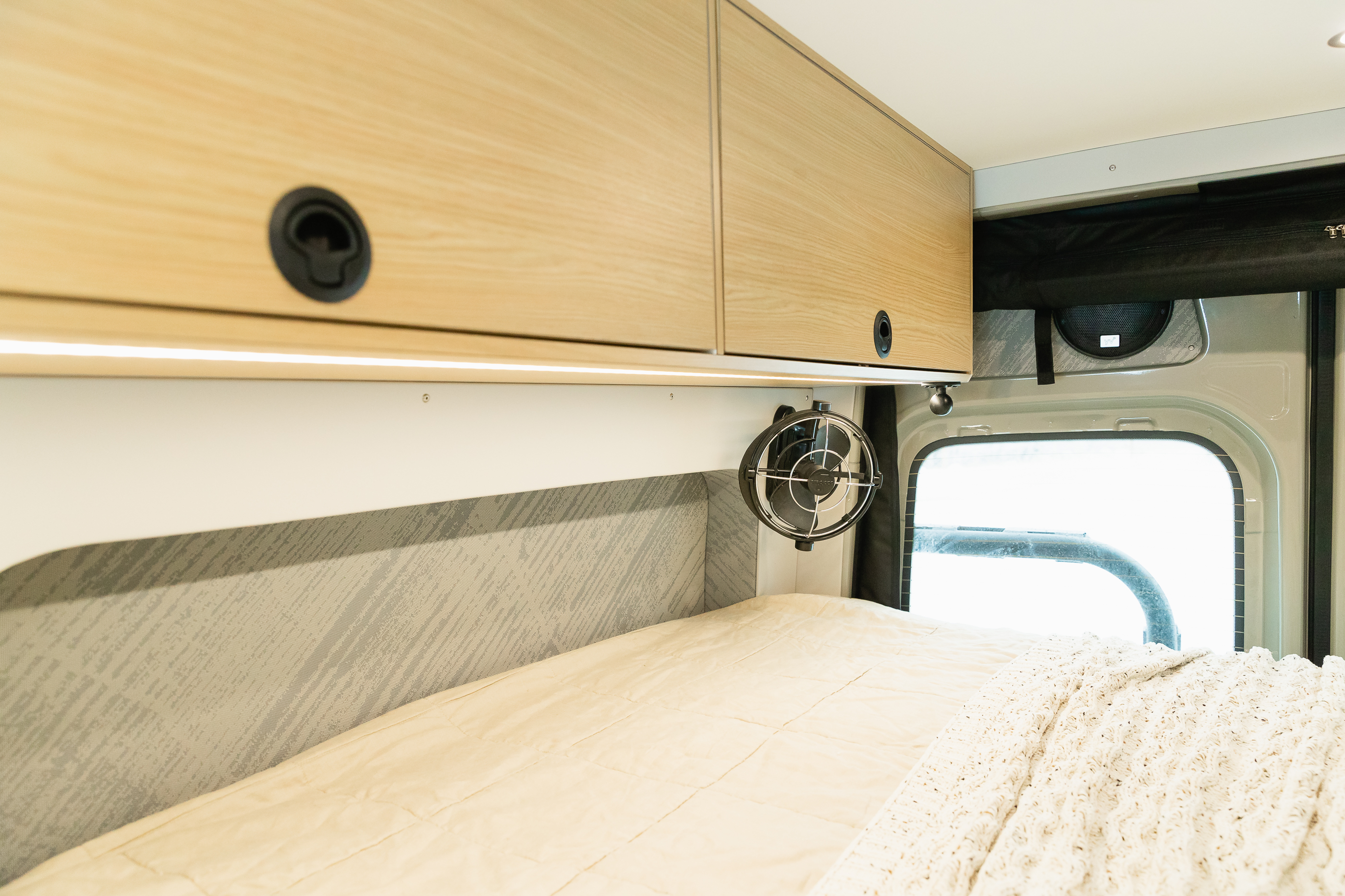 This RV rental interior features light wood cabinets, a cozy bed with cream bedding, and a small fan.. Mercedes-Benz Sprinter 2022