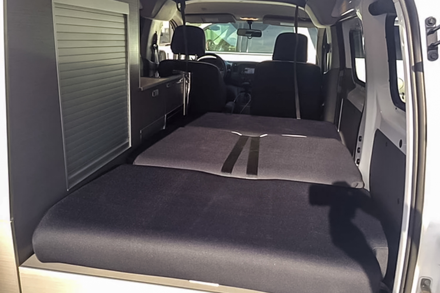Inside an RV rental: a flexible dark grey bed system unfolds towards the front seats, with sleek storage cabinets on the left.. Nissan Other 2021