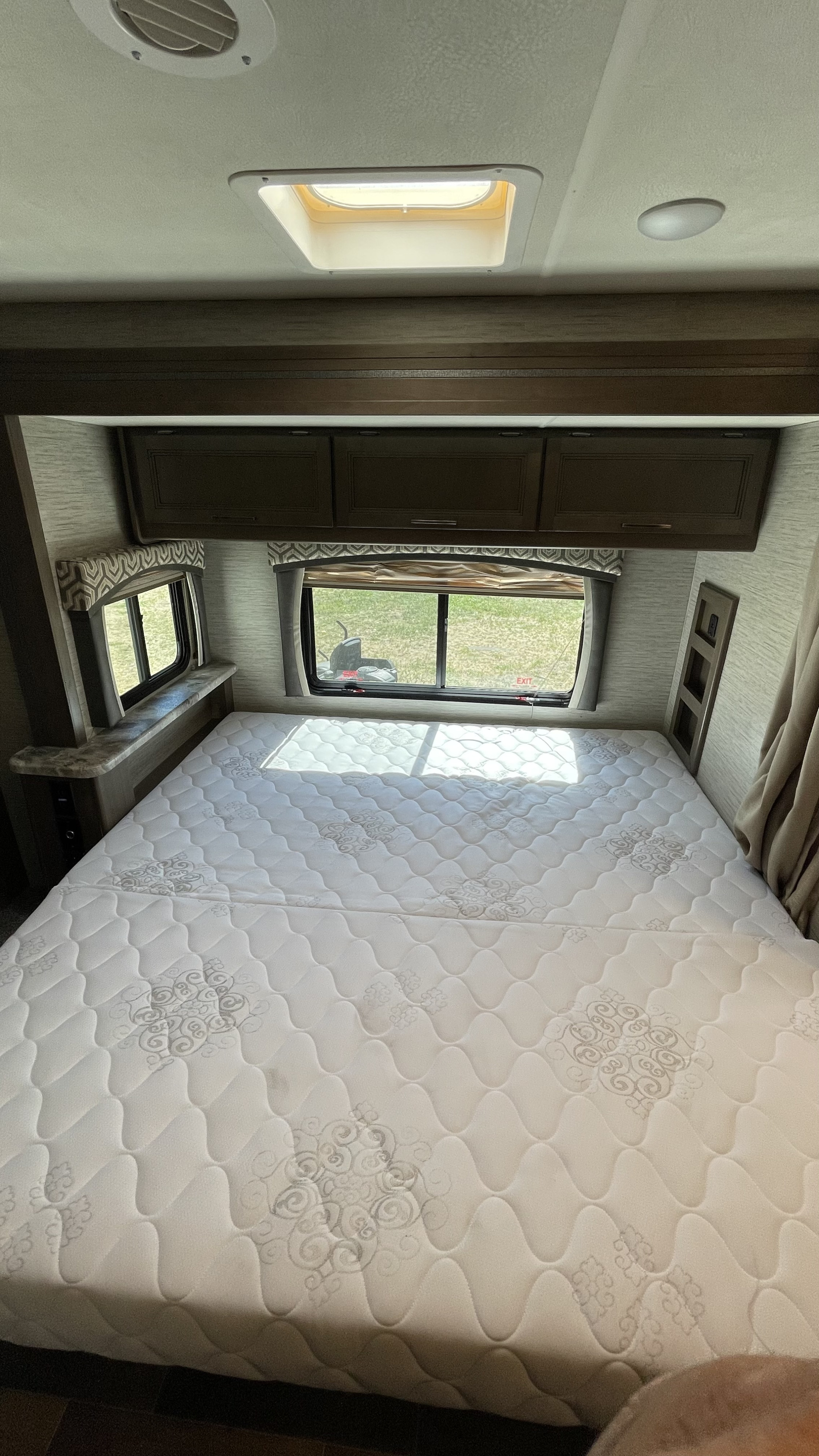 A bright RV rental bedroom with a white patterned mattress, multiple windows, overhead cabinets, and ceiling skylight.. Thor Chateau 2021