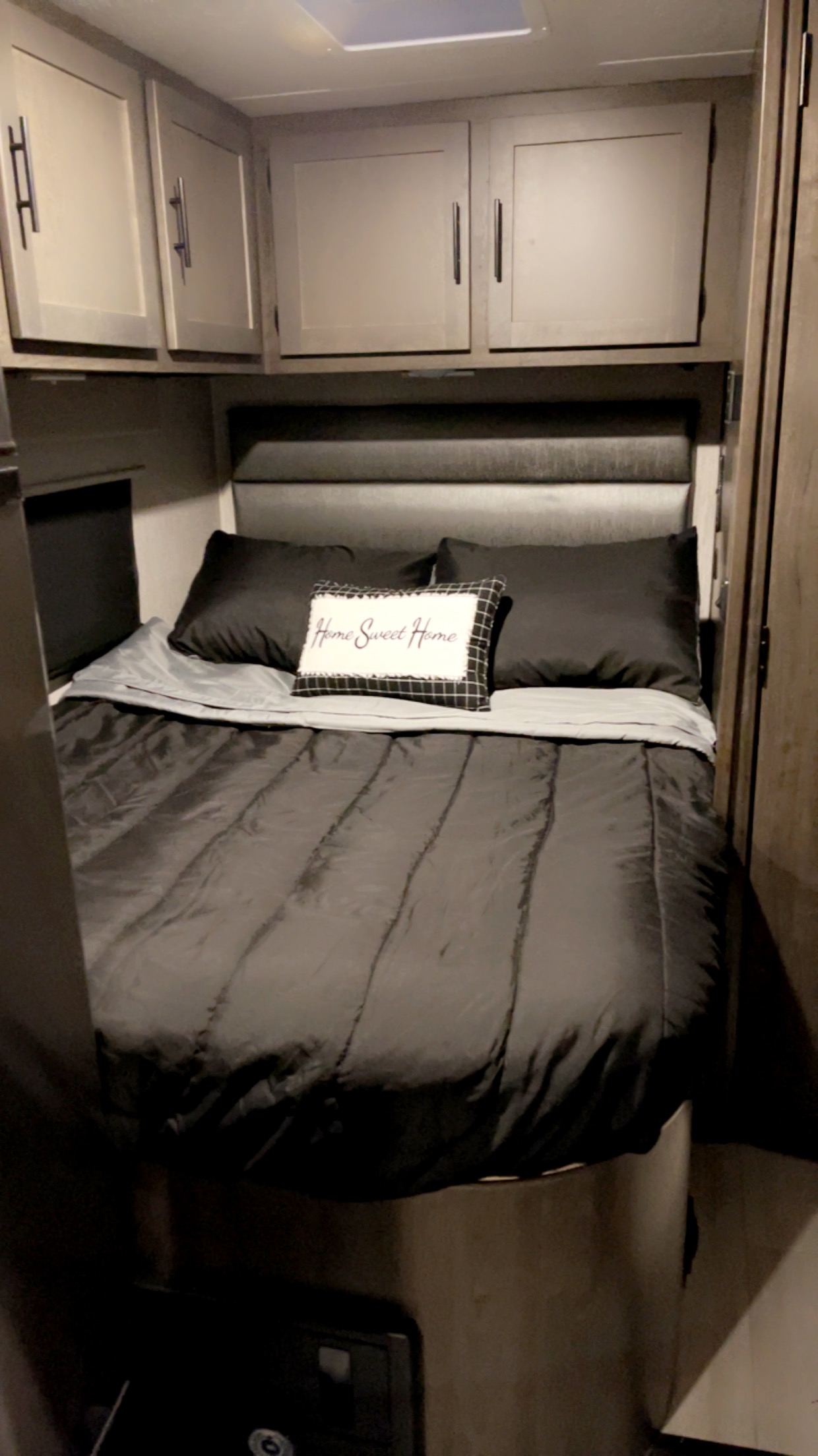 Inside this RV rental bedroom: a cozy space with a bed, dark bedding, "Home Sweet Home" pillow, and overhead cabinets.. Coachmen Prism 2023