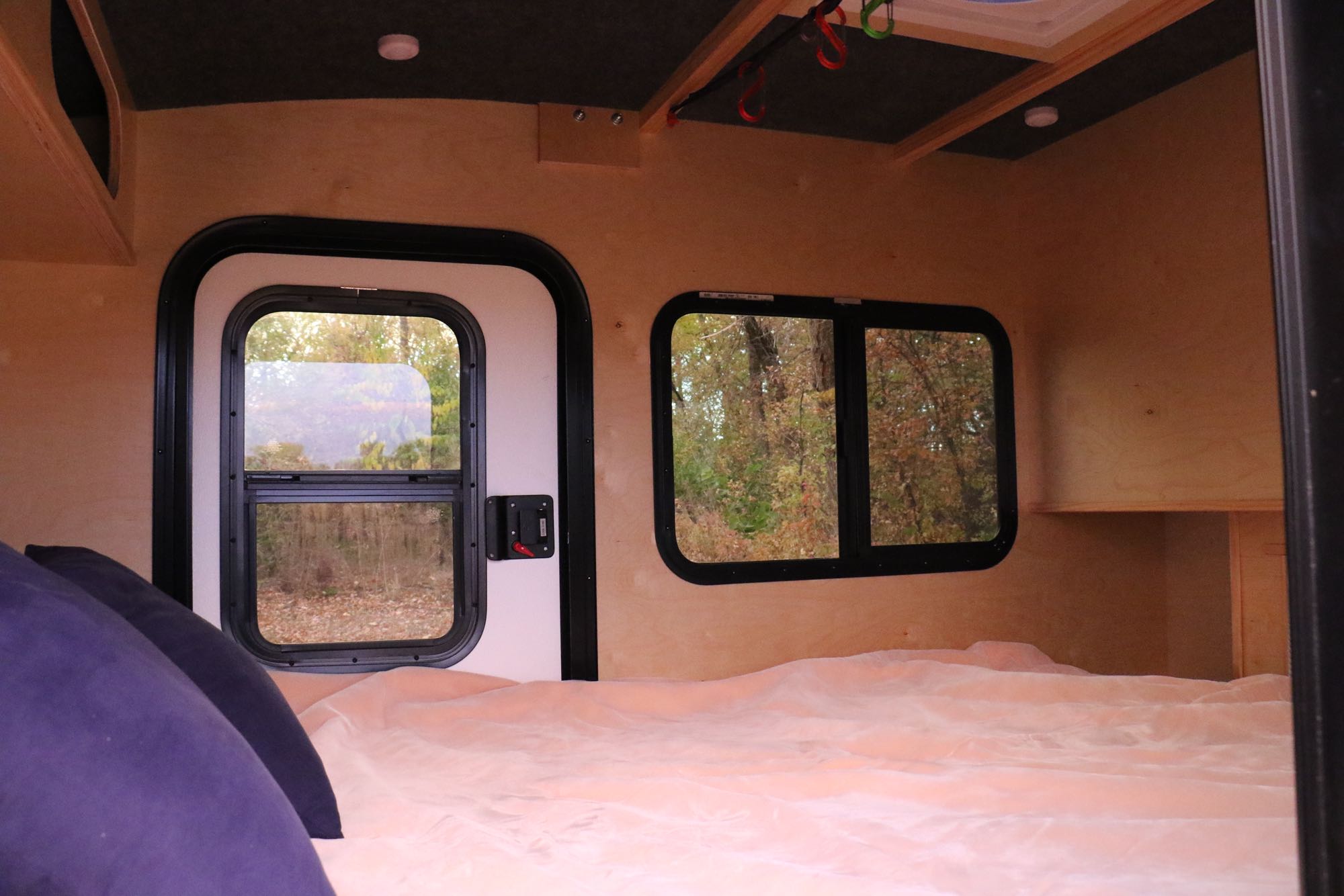Cozy RV rental interior: a bed with pink blanket, wood walls, and windows looking out to autumn trees. Camper living.. Outbound Extreme 2021