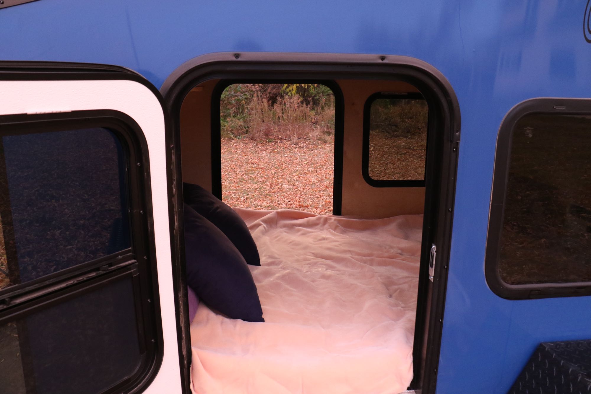 Inside a blue rv rental: a cozy bed with pillows looks out to a scene of autumn leaves on the ground.. Outbound Extreme 2021