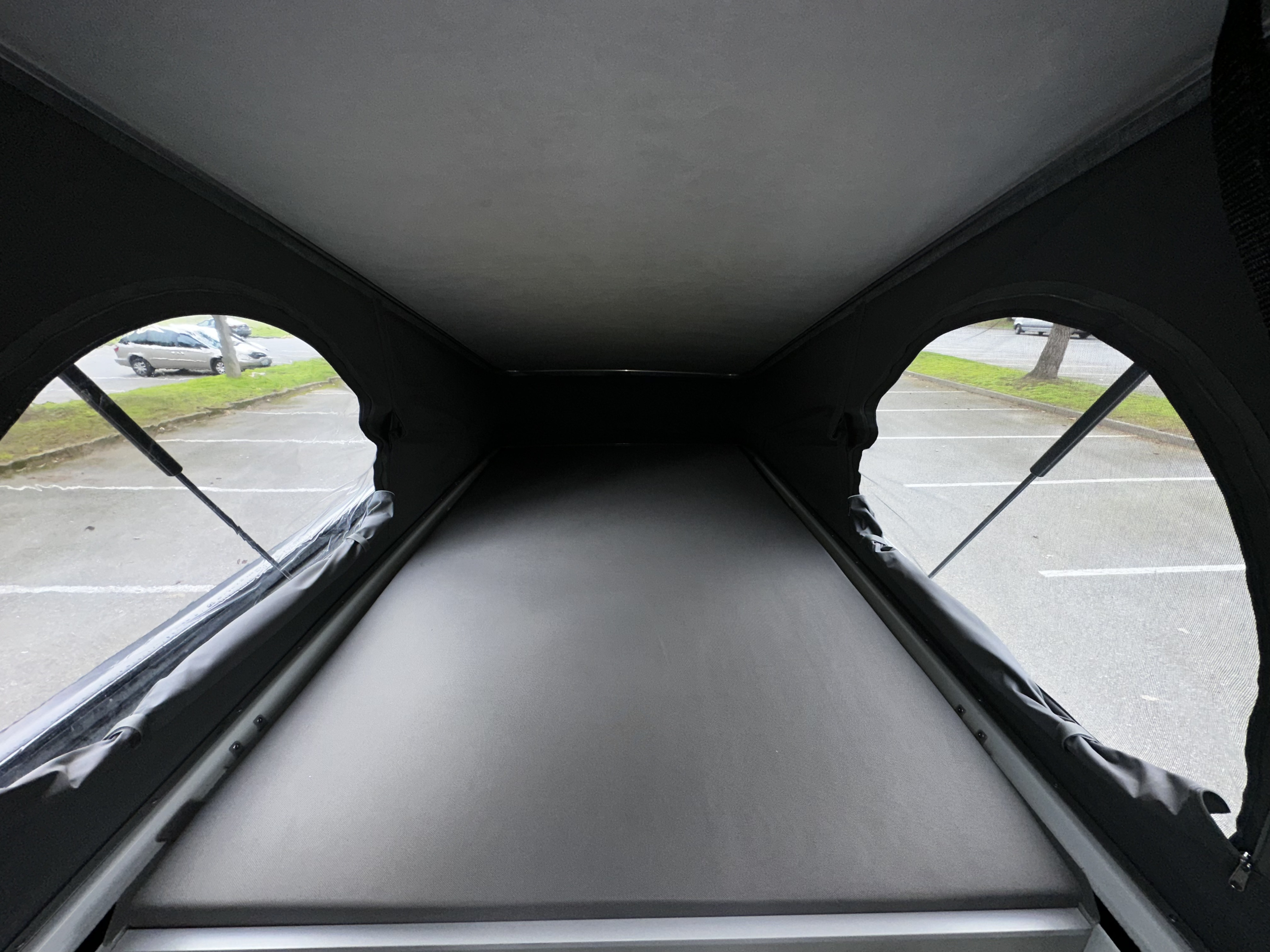 Inside an RV rental pop-top, showing the bed, side windows, and supports, with a parking lot visible outside.. Mercedes-Benz Metris 2016