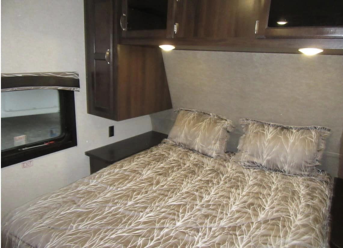 Inside an RV rental: cozy bedroom with patterned bedding, overhead cabinets, and a window with an 'EXIT' sign for travel comfort.. Jayco Jay Flight 2018