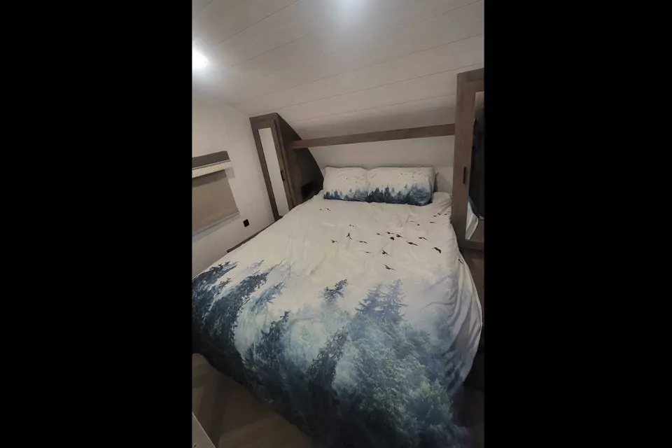 Serene RV rental bedroom with a comfortable bed featuring forest landscape bedding and built-in storage.. Forest River Wildwood X-Lite 2022