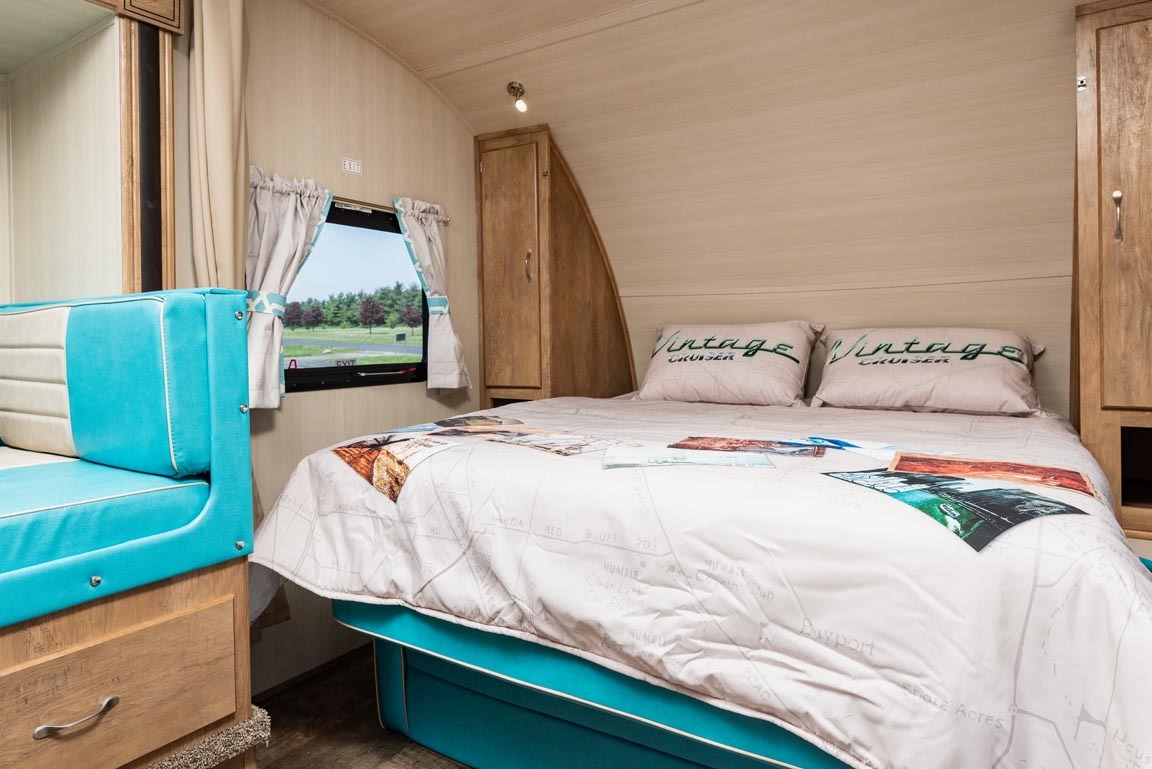 Inside an RV rental, a comfy bedroom features a teal bed, map-themed duvet, wooden cabinets, and an adjacent teal dinette.. Gulf Stream 23BHS Vintage Cruiser 2023
