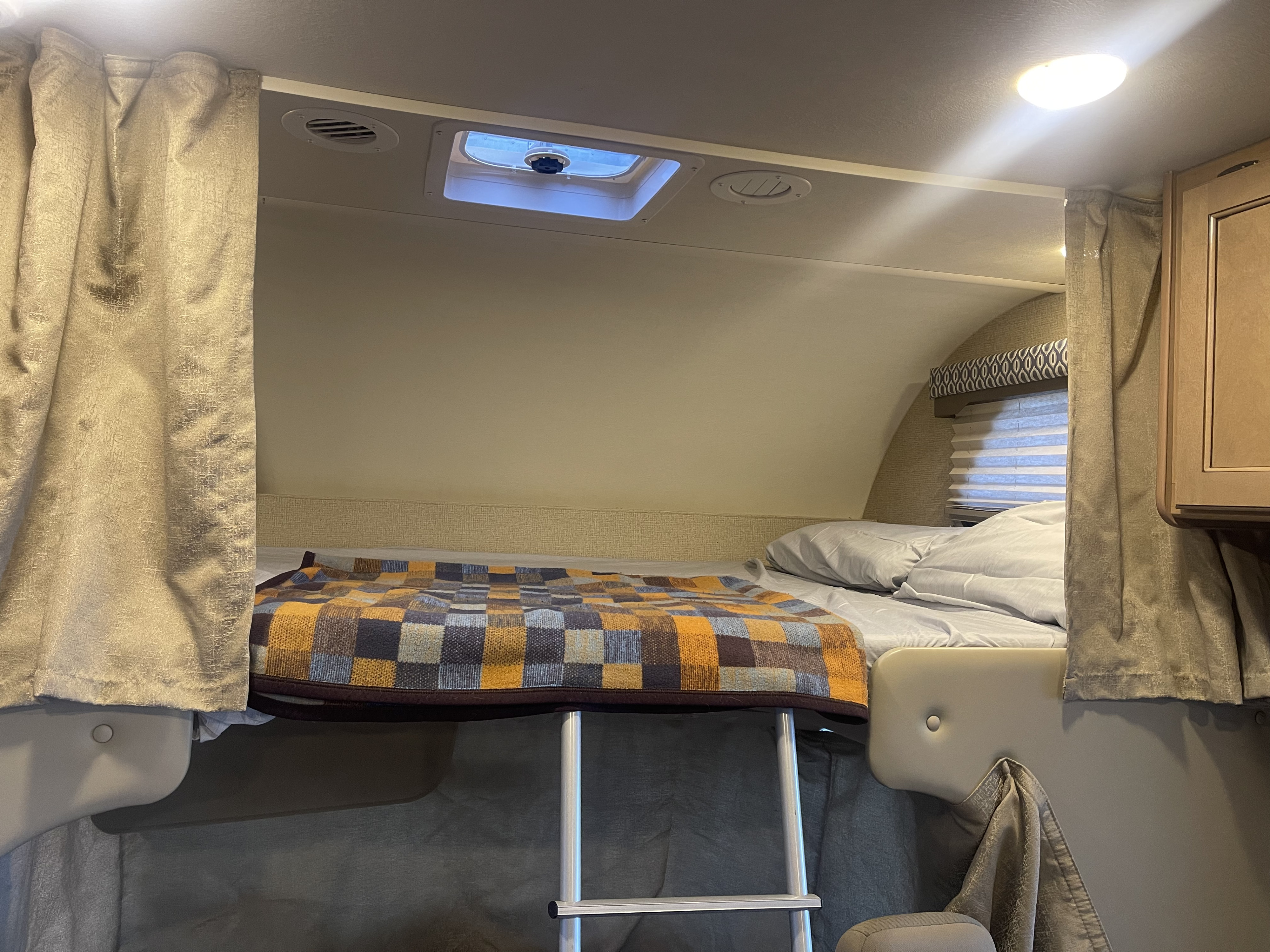 Cozy overhead bed in an RV rental, complete with a ladder, checkered blanket, and ceiling vent for comfortable mobile living.. Thor Freedom Elite 2019
