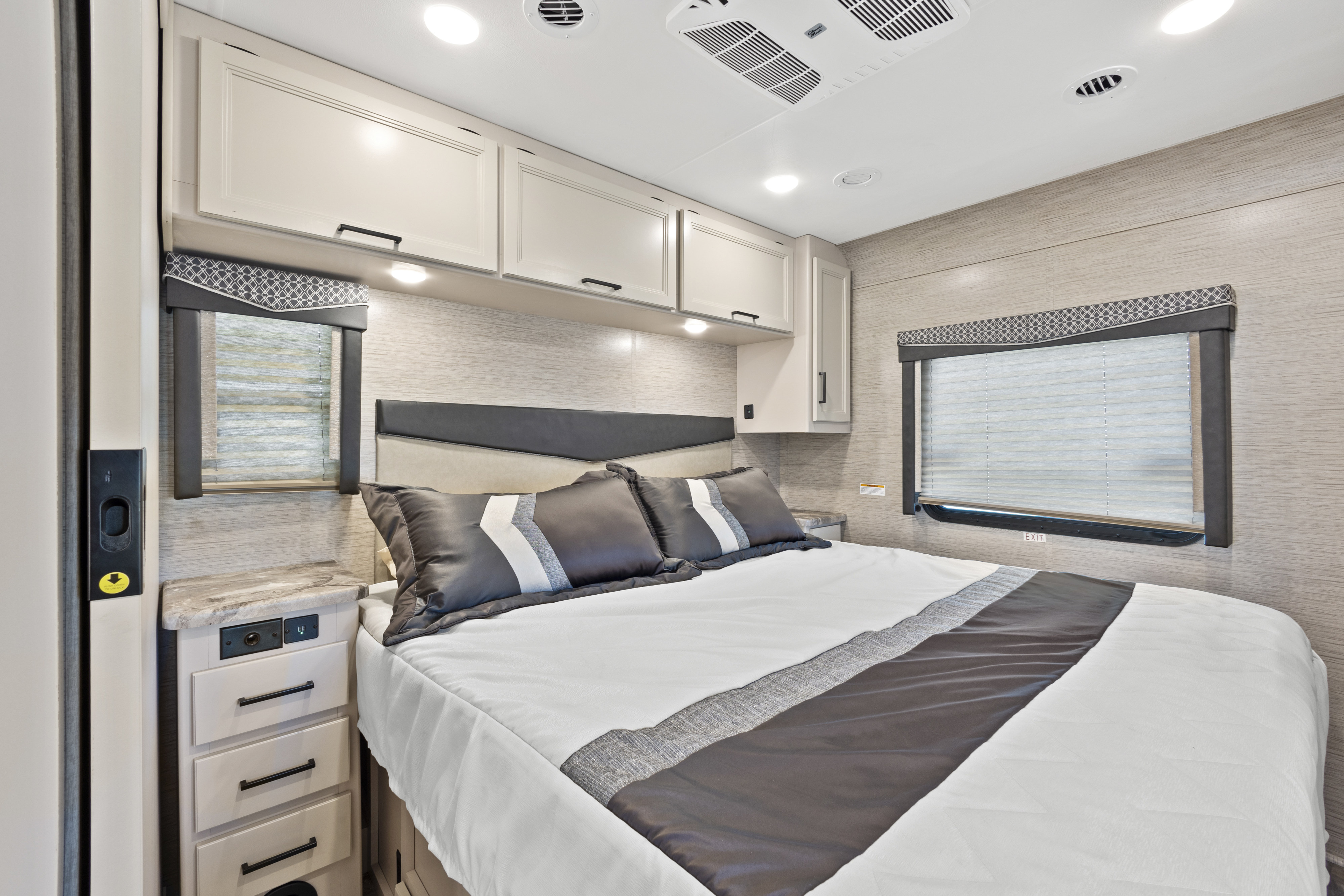 Cozy RV rental bedroom showcasing a comfortable bed with neutral decor, overhead storage cabinets, and dual windows.. Thor Motor Coach A.C.E 2023