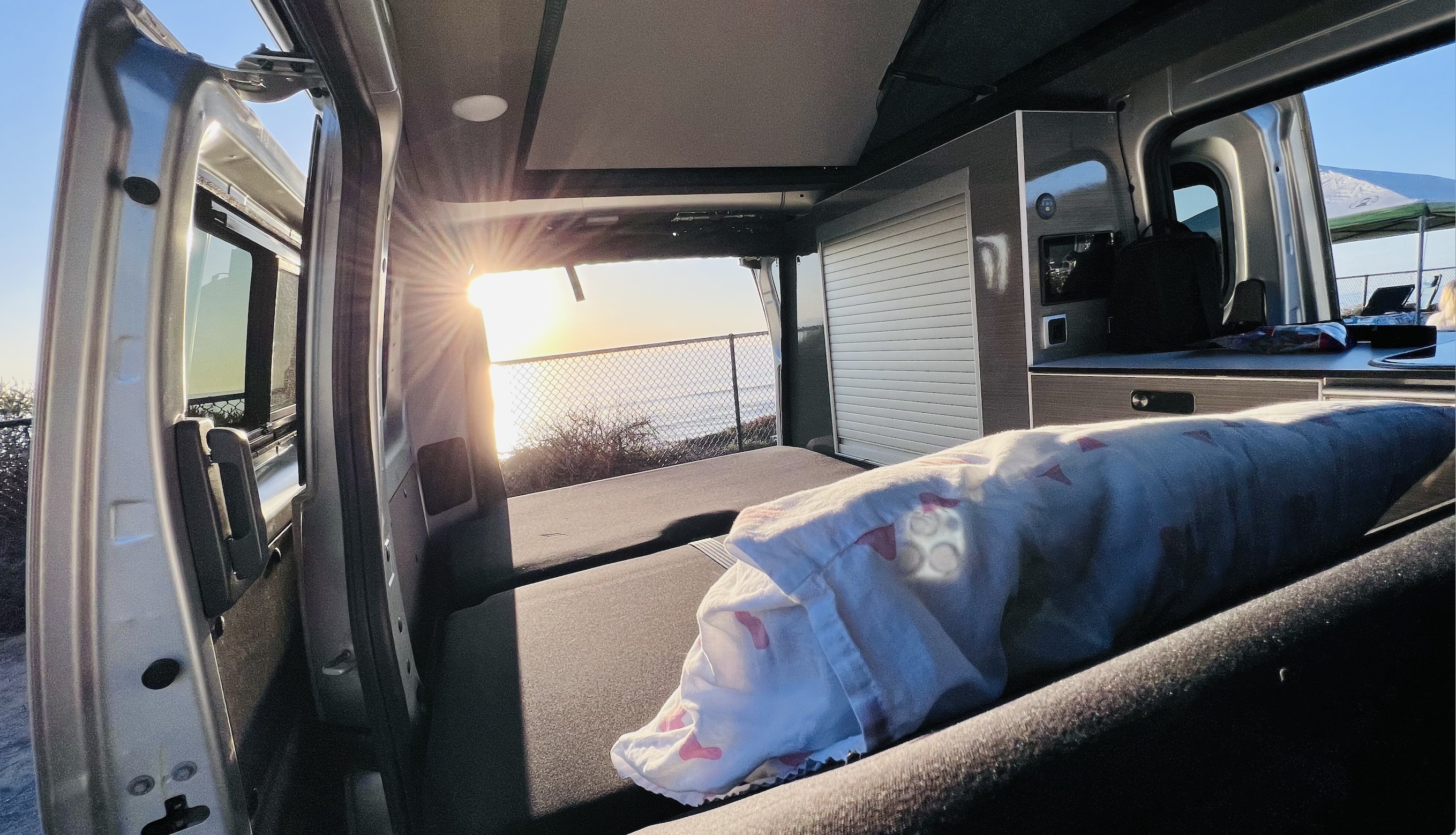 Bright rv rental interior at sunset. A bed and kitchen unit face open doors, revealing an ocean view and setting sun.. Nissan Other 2021