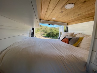 This inviting RV rental camper van features a cozy bed, white walls, wood ceiling, and scenic views from its open rear.. Ram Promaster 2021